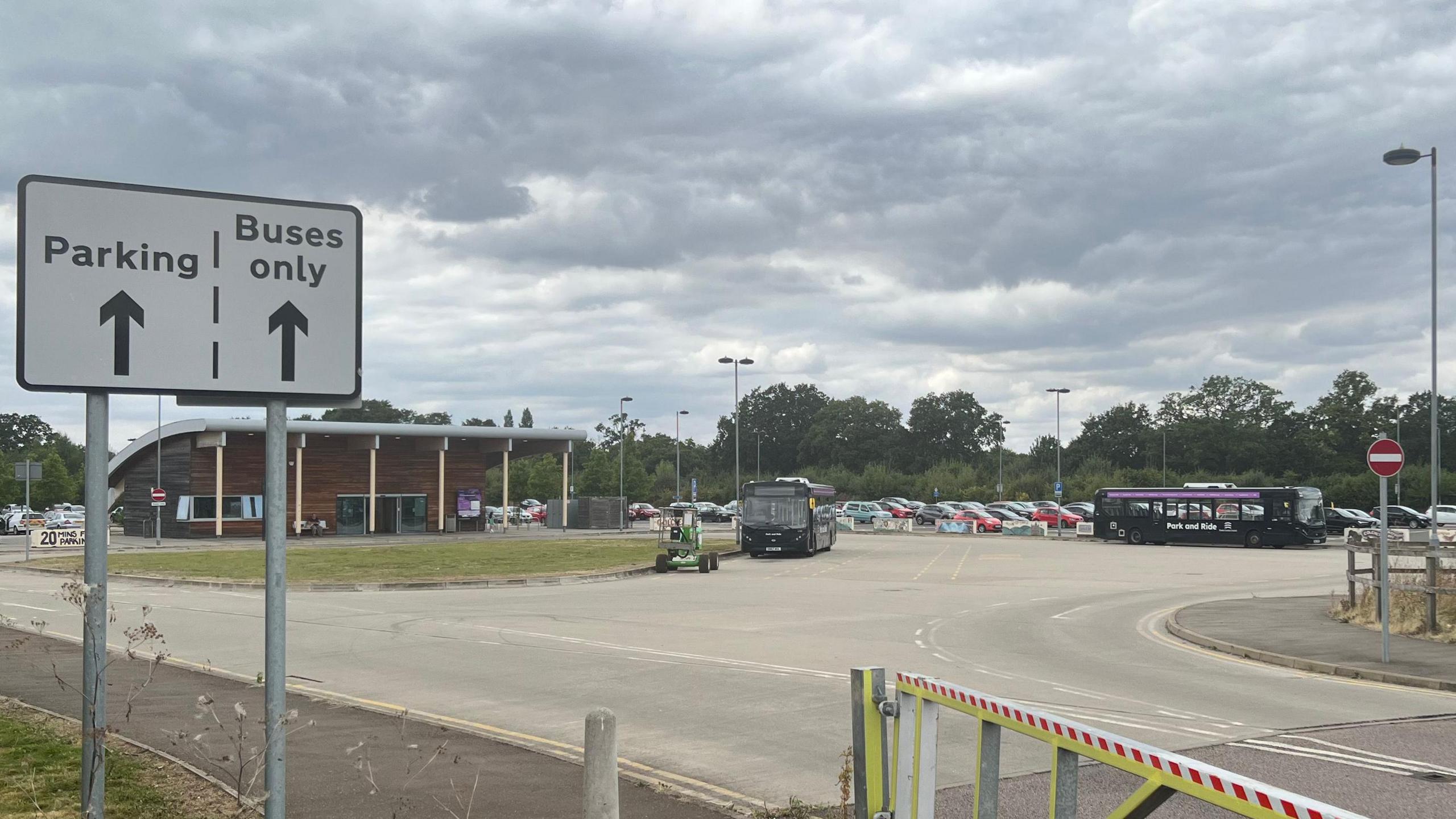 A view of the entrance to Colchester Park and Ride car park. A sign signals where vehicles can park and which direction is just for buses. Buses can be seen parked up in the distance with cars parked in the car park behind them.