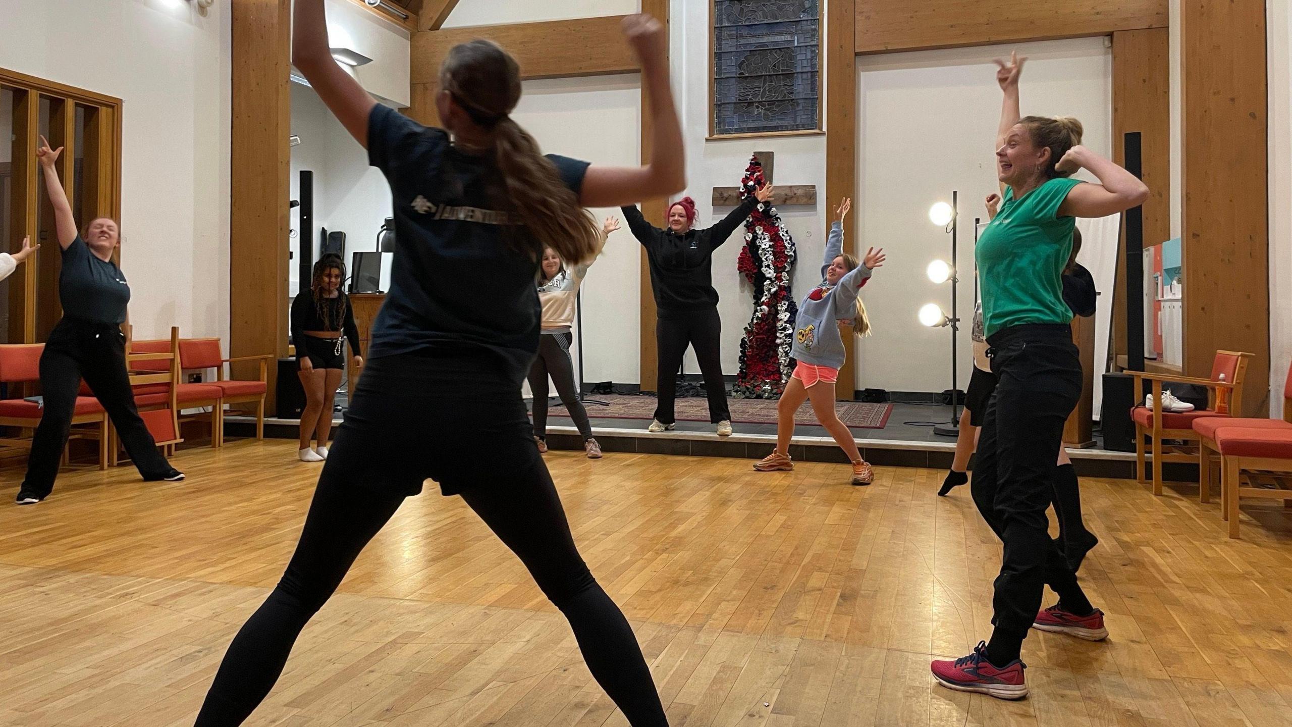 Ten girls and women stand in a circle in a hall with wooden floors and chairs to the side. They are standing with their legs apart and are stretching their arms in the air.