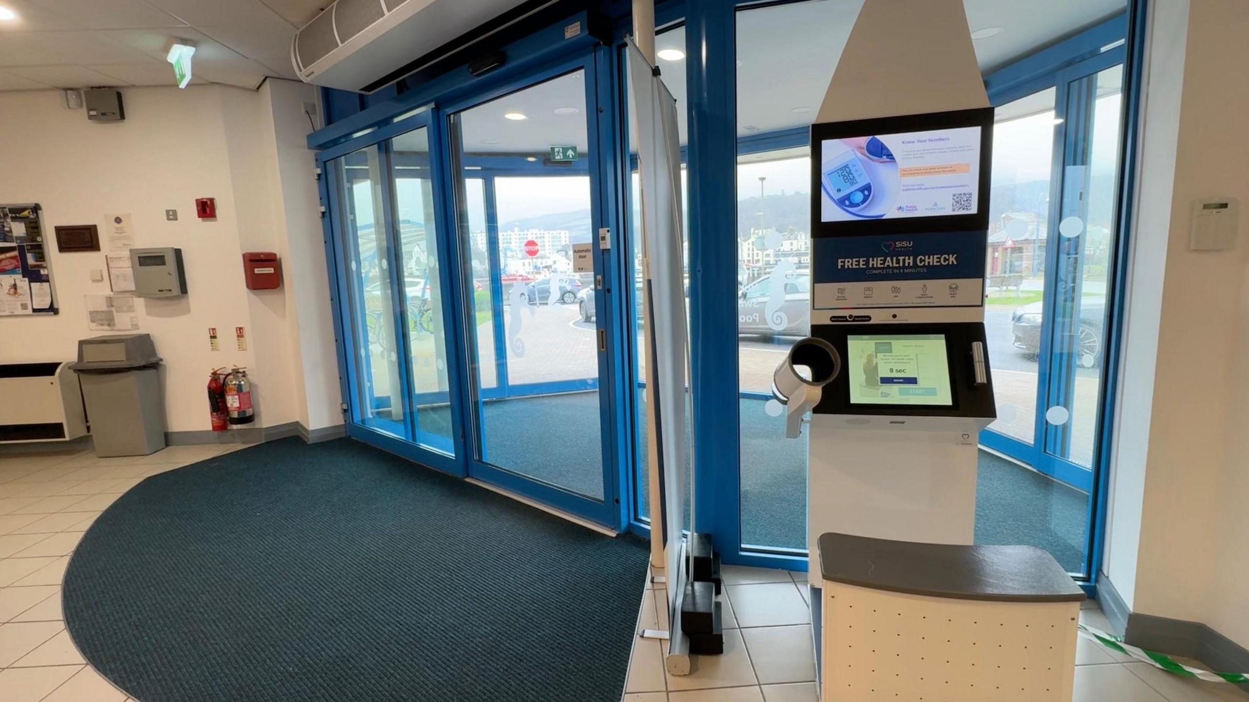 The tall white machine on the right of the foyer has two screens, one above the other, and a seat in front. To the left of the machine is a screen, and the blue automatic doors to the swimming pool entrance on the left with a large curved door mat.