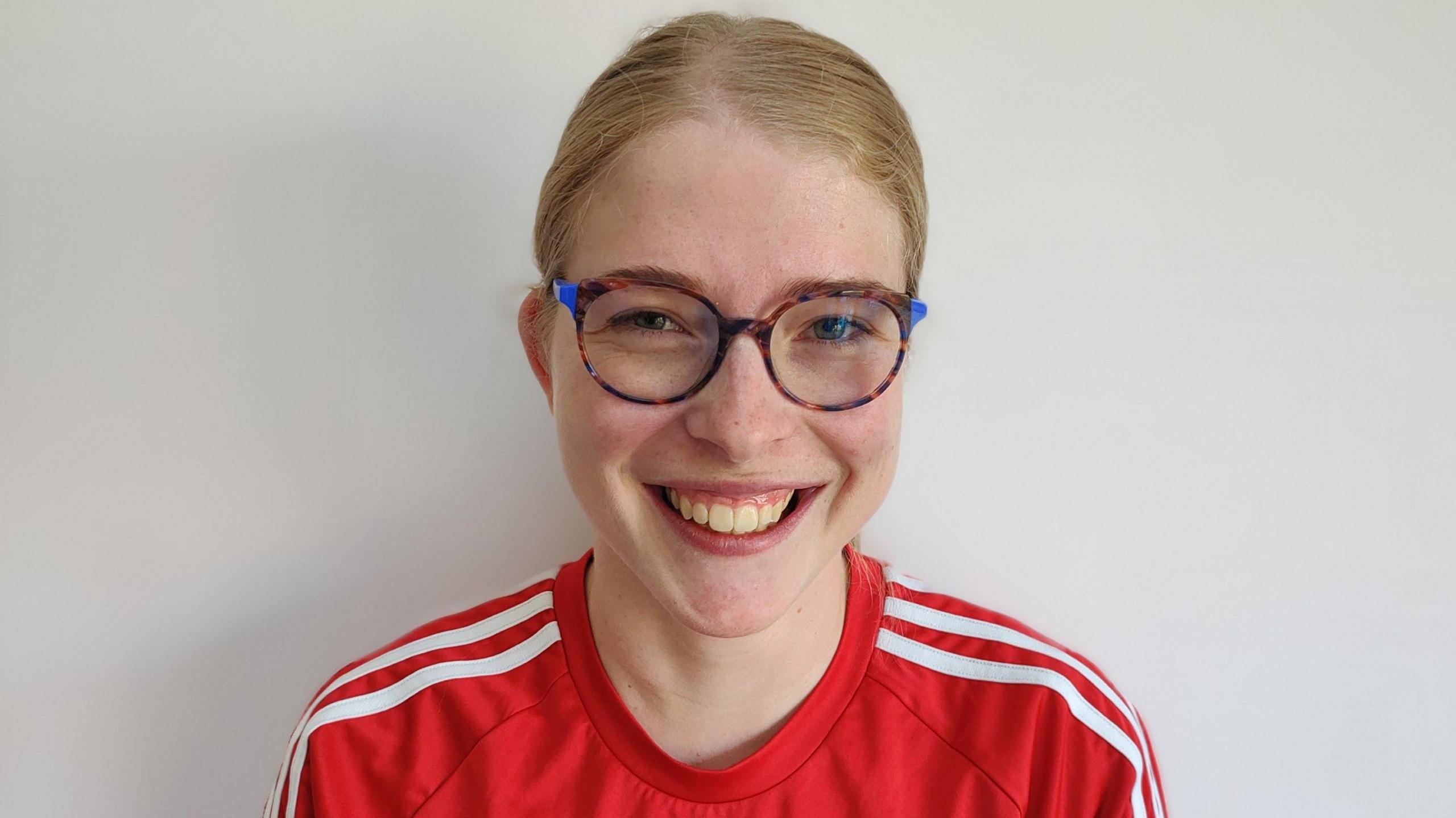 Jess Lowe, a 25-year-old white woman with strawberry blonde hair which is tied up. She is wearing a red Adidas T-shirt and glasses with a brown and blue frame. 