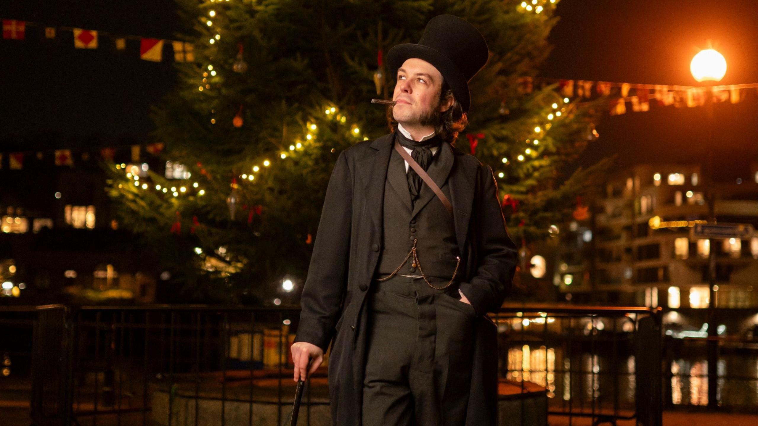 A man dressed as engineer Isambard Kingdom Brunel stands in front of a Christmas tree on the dockside in Bristol next to the SS Great Britain. The picture is taken at night and the tree's lights are illuminated. Behind the tree the flats on the other side of the harbour are visible