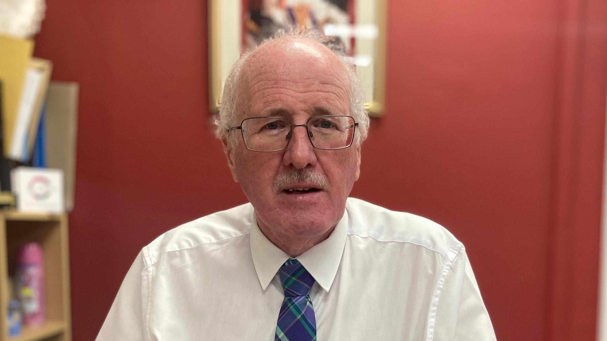 Jim Shannon. He is bald with white hair at the sides, wearing glasses, a white shirt and purple and green tartan tie. In the background there is a bookcase and artwork on a red wall.