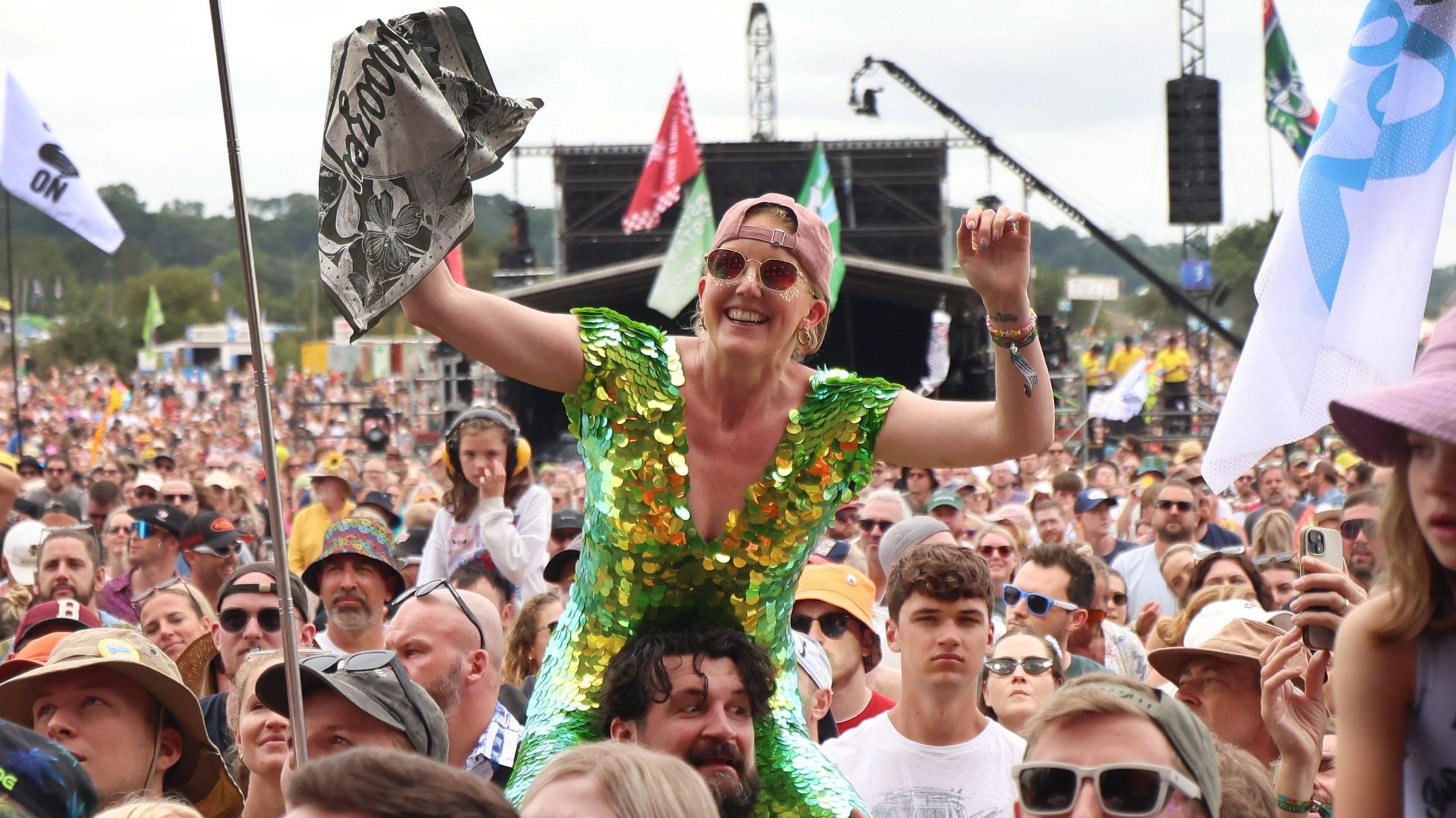 A woman wearing a green and yellow sequin dress cheers with her arms in the air as she sits on a man's shoulders in the crowd. There are people and flags all around her.