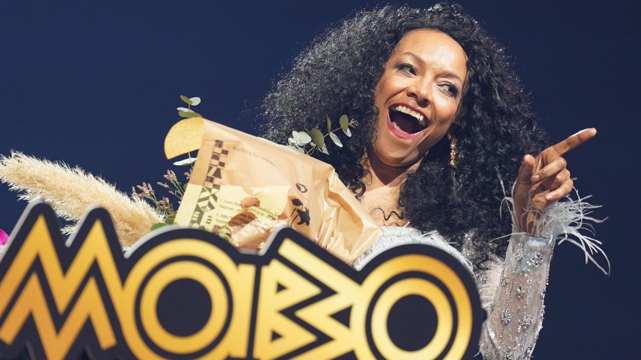 Kanya King on stage after being recognised with a surprise paving the way award. She holds a bouquet of flowers while wearing a feathered silver dress