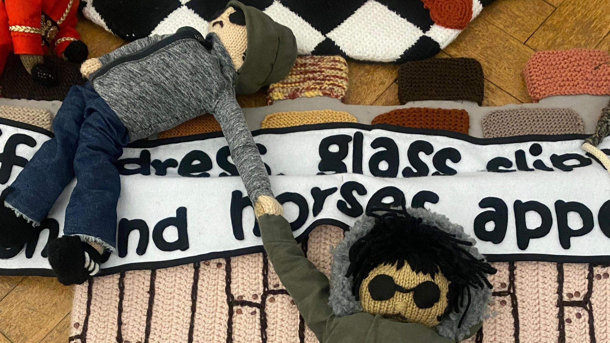 Two characters, made out of wool, that look like Liam and Noel Gallagher. They are holding on to each other. One has sunglasses on with a coat and hood. The other is wearing a hat with dark clothing. They are beside a banner. 