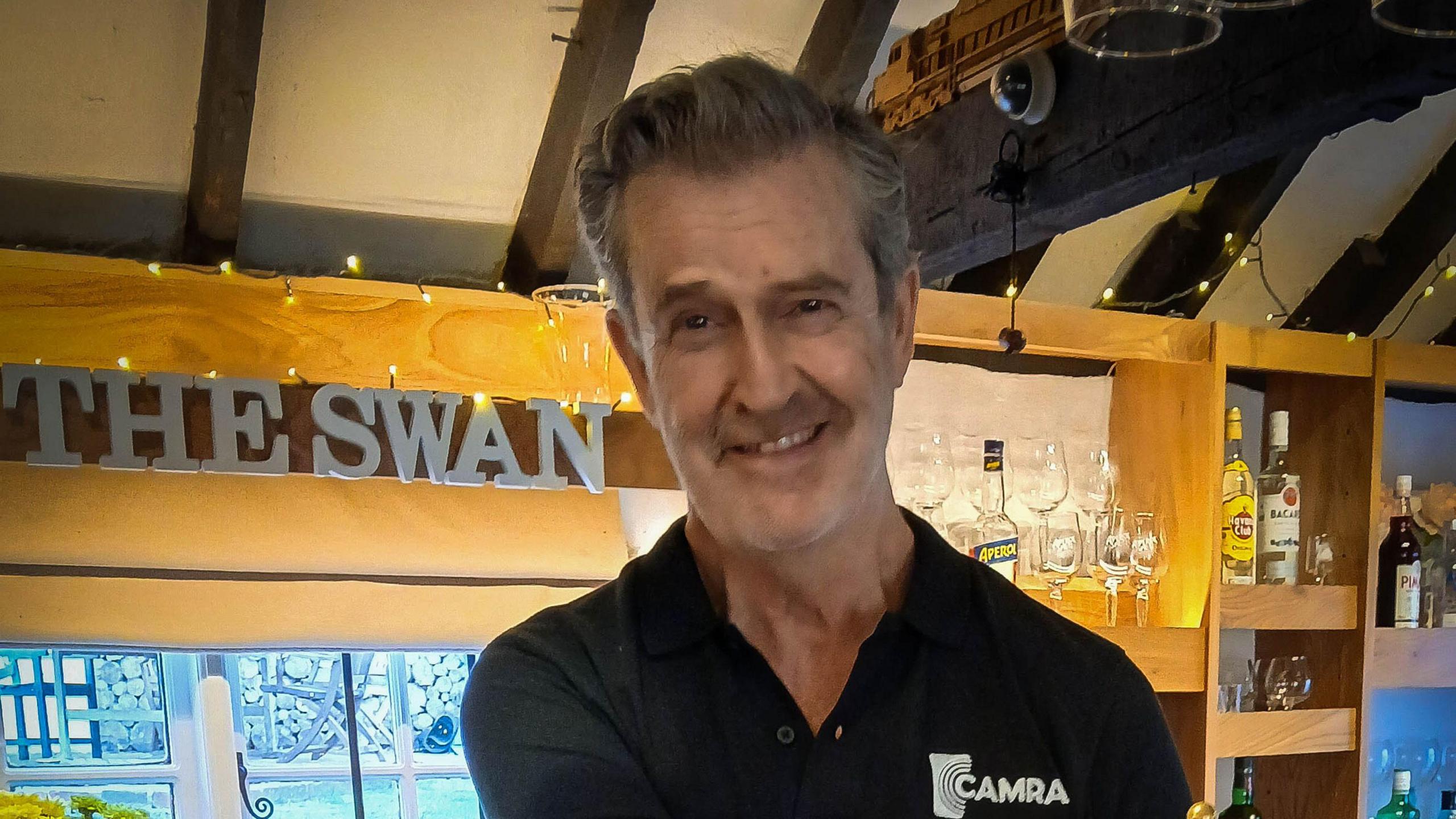 The actor Rupert Everett, wearing a shirt with the word CAMRA written on it, smiling at the camera while standing behind the bar in a pub.