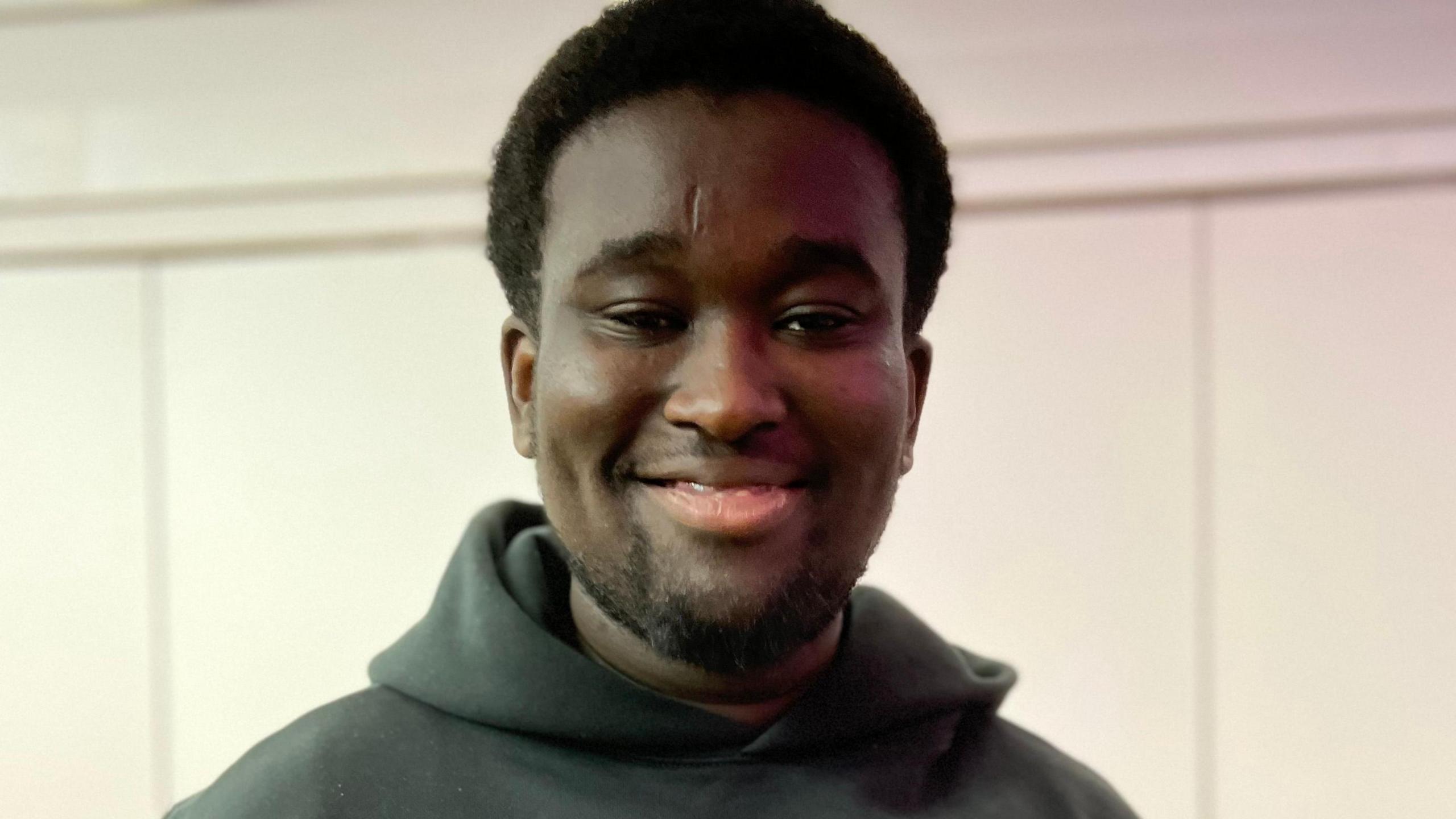 A young man stands with his arms clasped behind him. He is smiling. He wears a charcoal coloured sweatshirt.