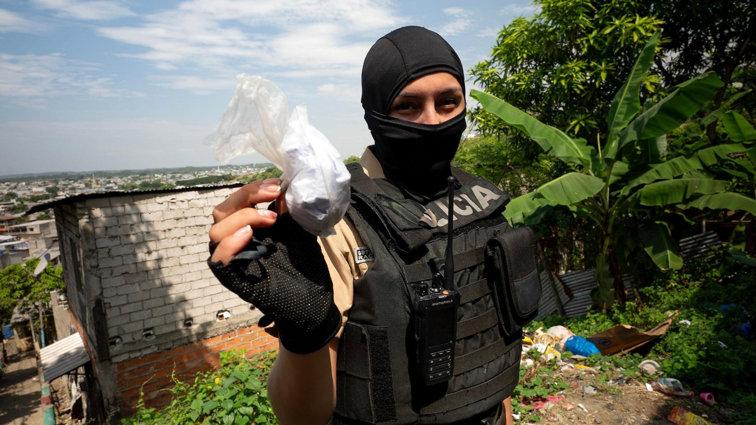 A police officer in a balaclava and body armour holds up a plastic bag filled with white powder.