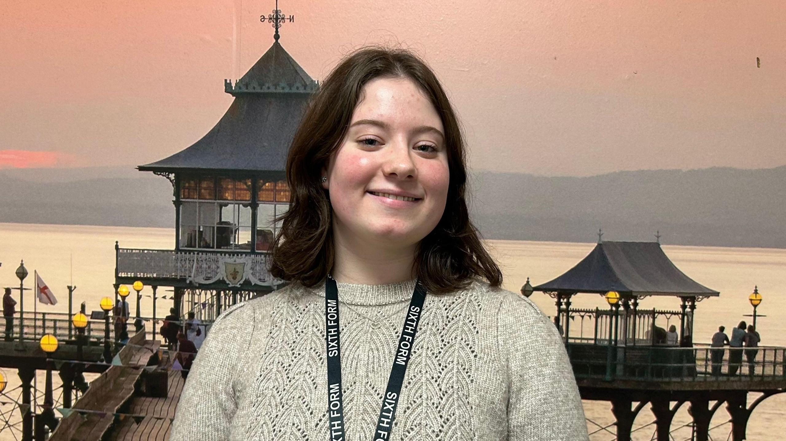 Clara, who has shoulder-length dark hair, is smiling and looking at the camera. She is wearing a light grey knitted jumper and a sixth form black lanyard. Behind her is a picture of Clevedon Pier.