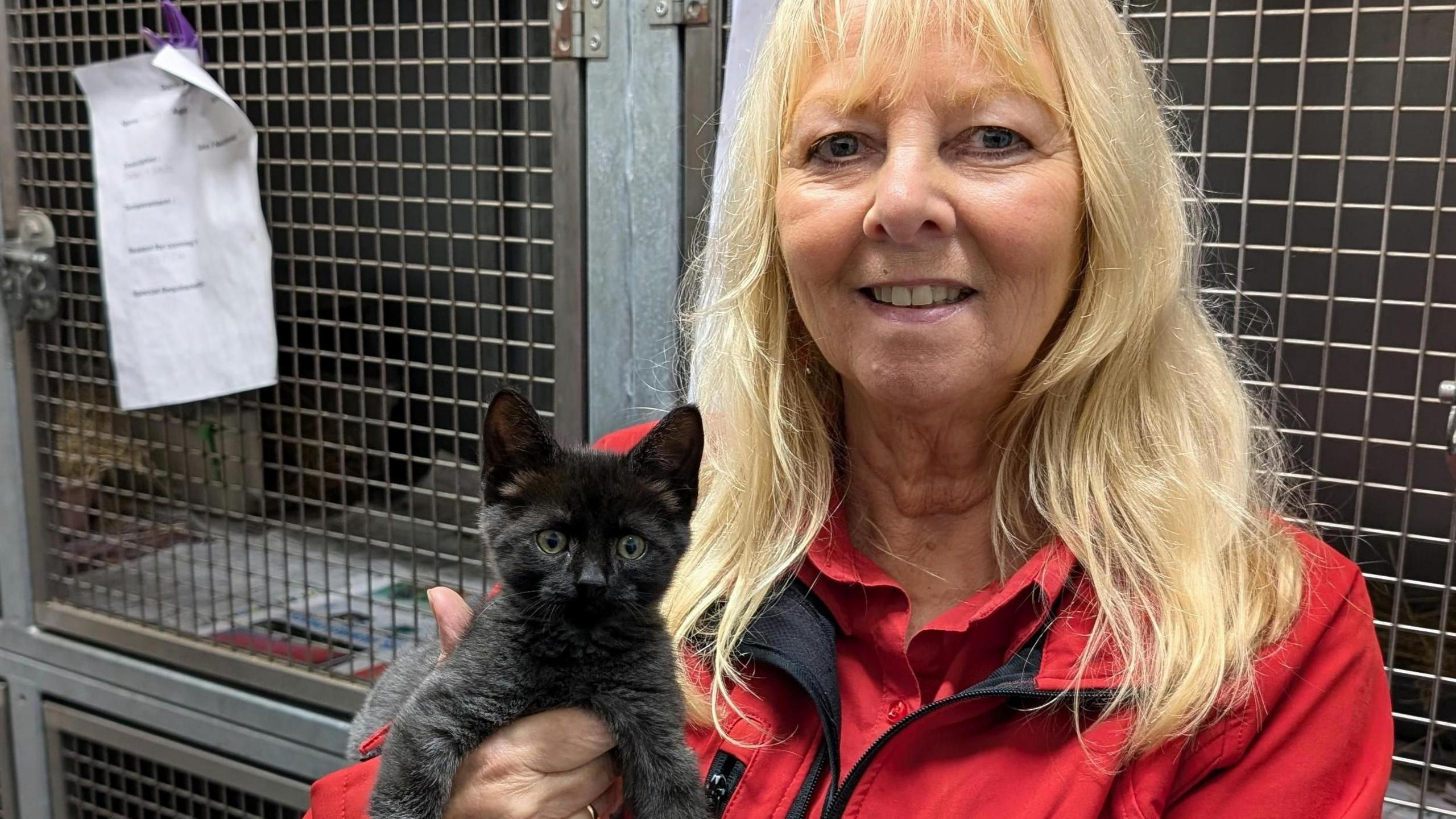 A woman with long blonde hair wearing a red jacked with the Woodside logo. She is holding a black kitten and both are looking directly into the camera. 