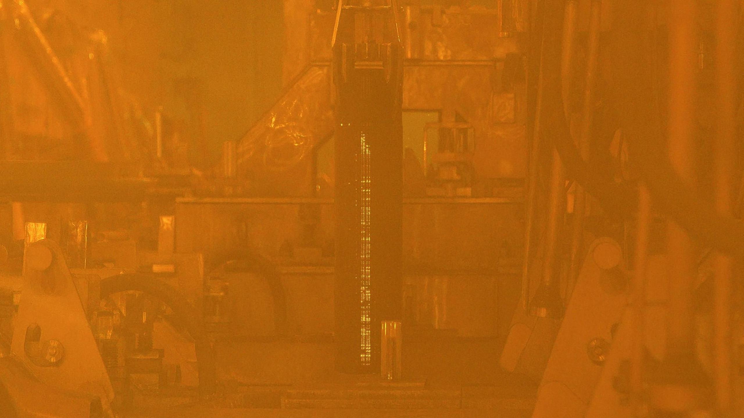 Plutonium is a product of reprocessing spent nuclear fuel, seen here from behind lead-lined glass. A rod of nuclear fuel is extracted, by a robotic arm, from a container. The thick, protective lead-lined glass gives a yellow tint to the scene and the nuclear fuel rod is glows because it is highly radioactive.   