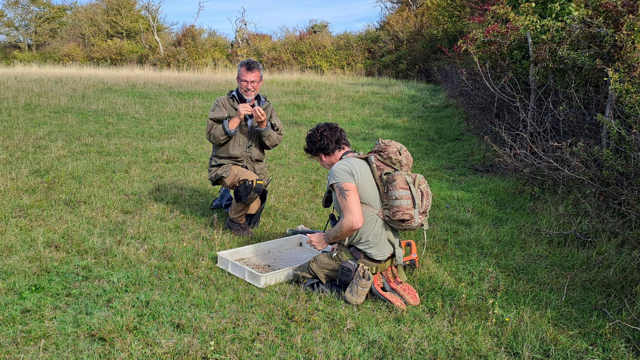 Two men sitting on grass, looking at something in a plastic tray