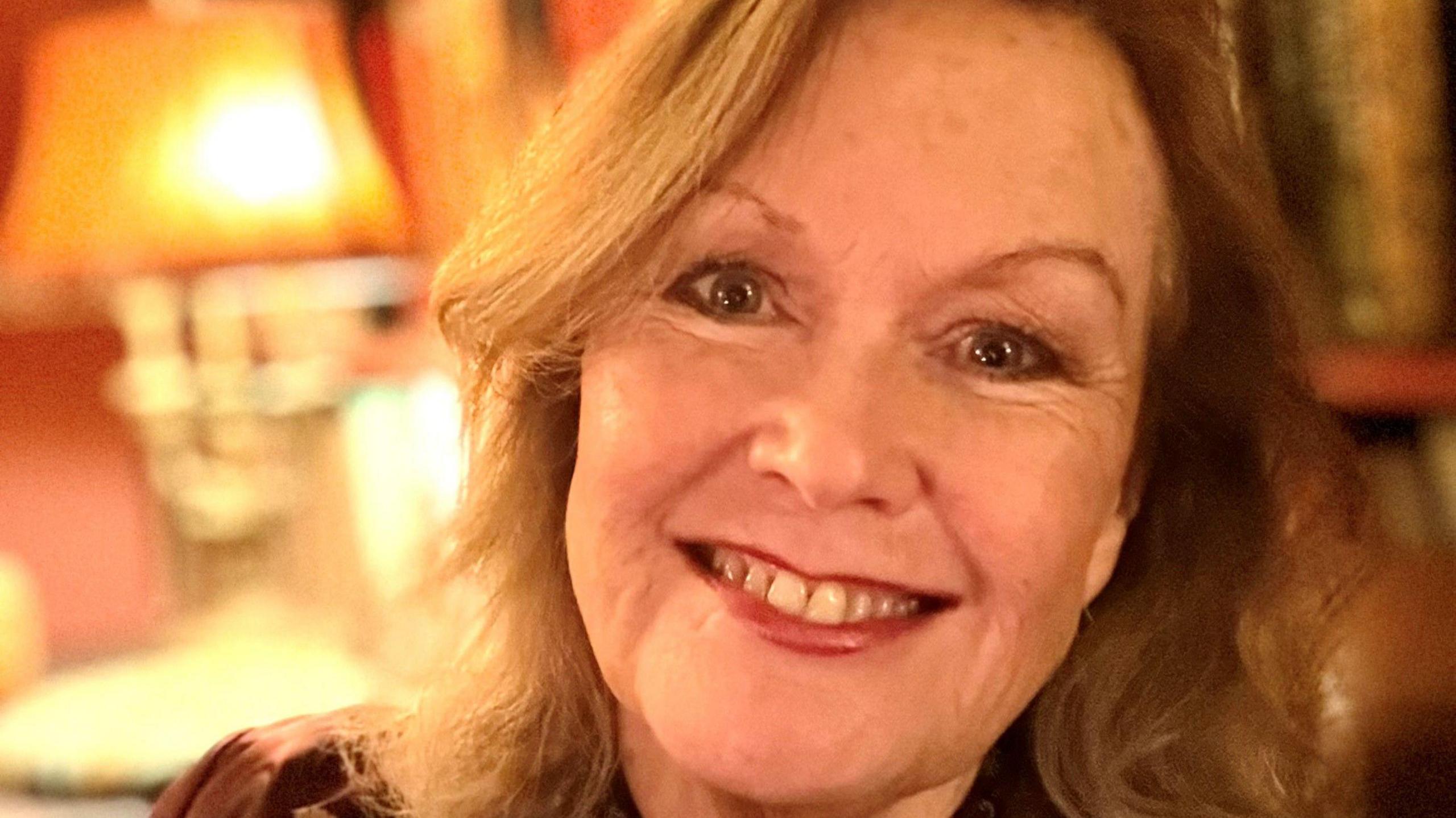 A close up photo of Wendy Holden. She smiles at the camera. She has shoulder length strawberry blonde hair that is slightly curled.