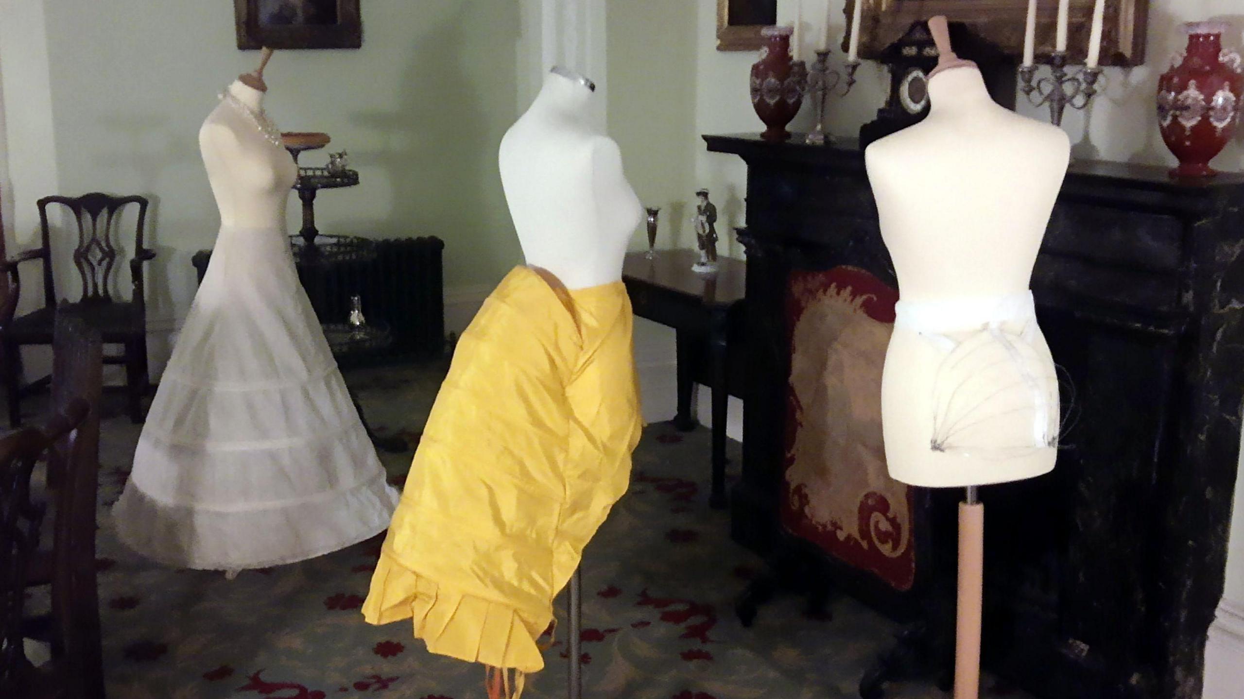 A historical fashion exhibit with three dresses in a vintage-style room. The first mannequin on the left wears a long, white layered and hooped skirt. The mannequin in the middle has a bright yellow skirt with ruffles on. The third mannequin on the right has a small, white undergarment on. In the background there are chairs and tables, as well as decorative items like candlesticks and vases on top of a fireplace.