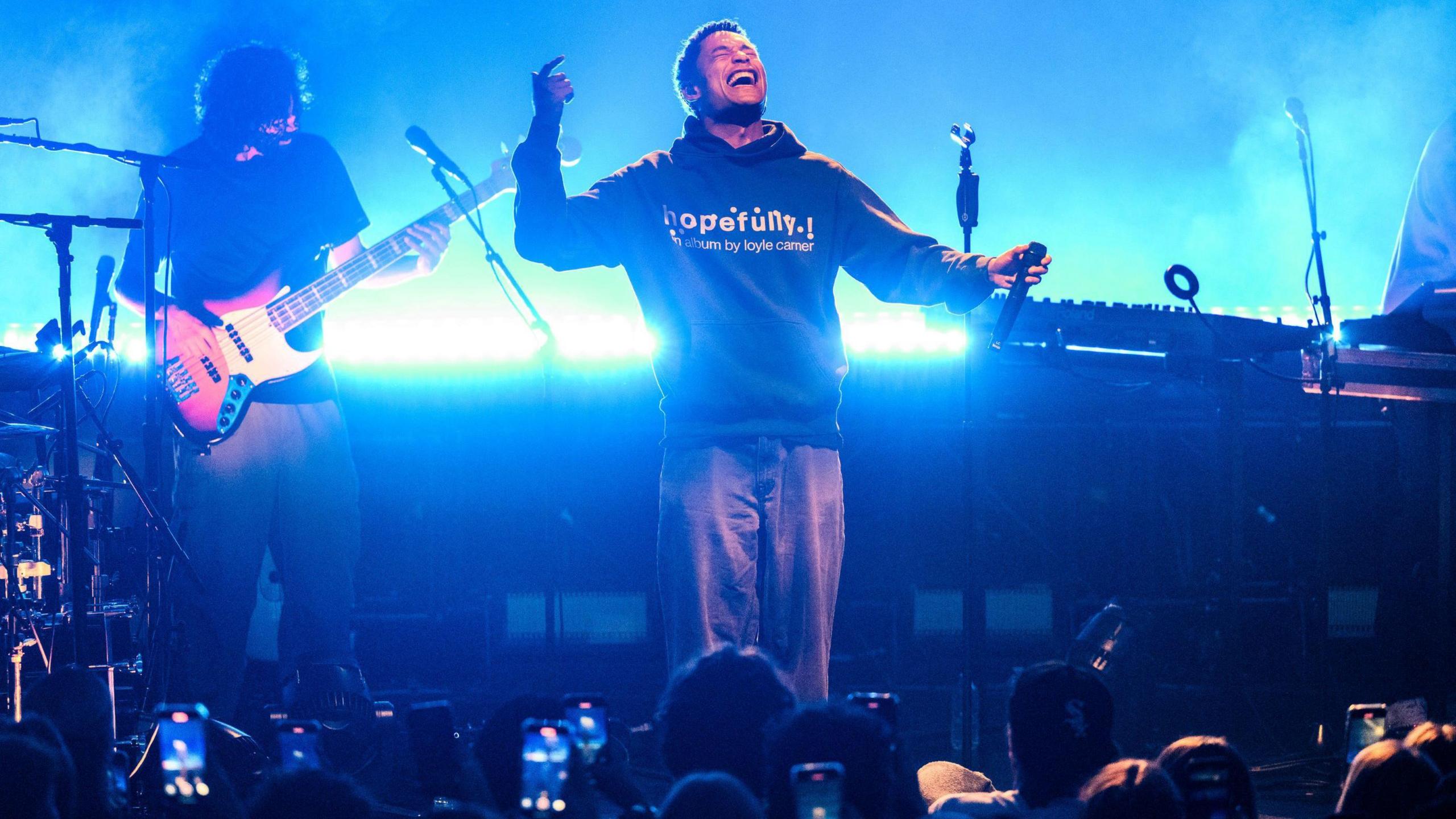Loyle Carner, wearing a hoodie and jeans, performs on stage at the O2 Academy in Bristol, with other musicians visible behind him.