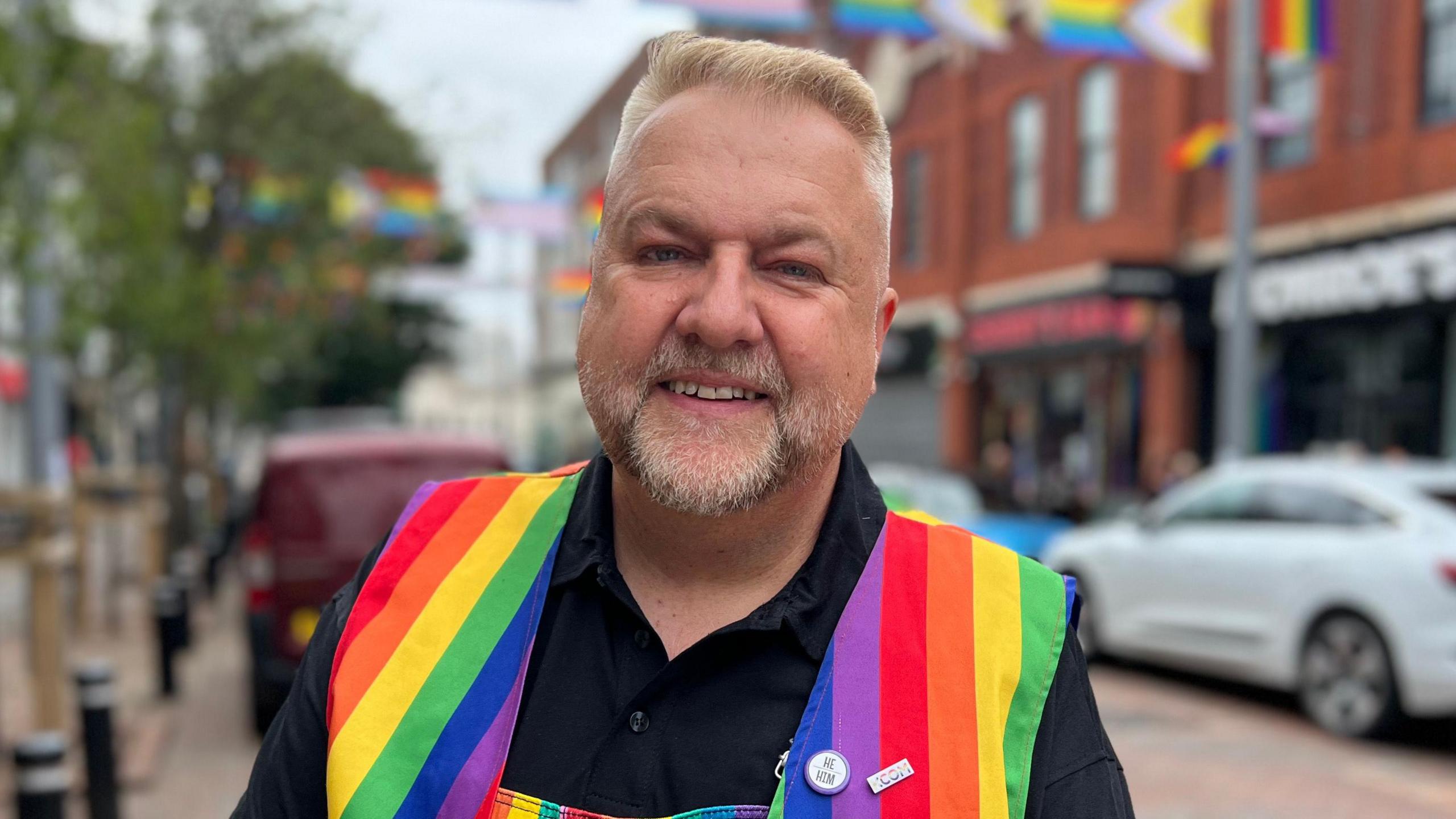 A man is stood in a street. He is wearing a black long sleeved shirt and a rainbow-patterned apron. He has short blond hair and a short grey beard. 
