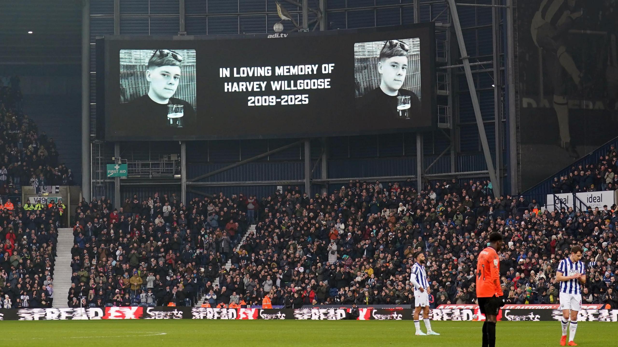 A screen at The Hawthorns football stadium reading 'in loving memory of Harvey Willgoose 2009-2025' with fans beneath it. Three players can be seen on the pitch.