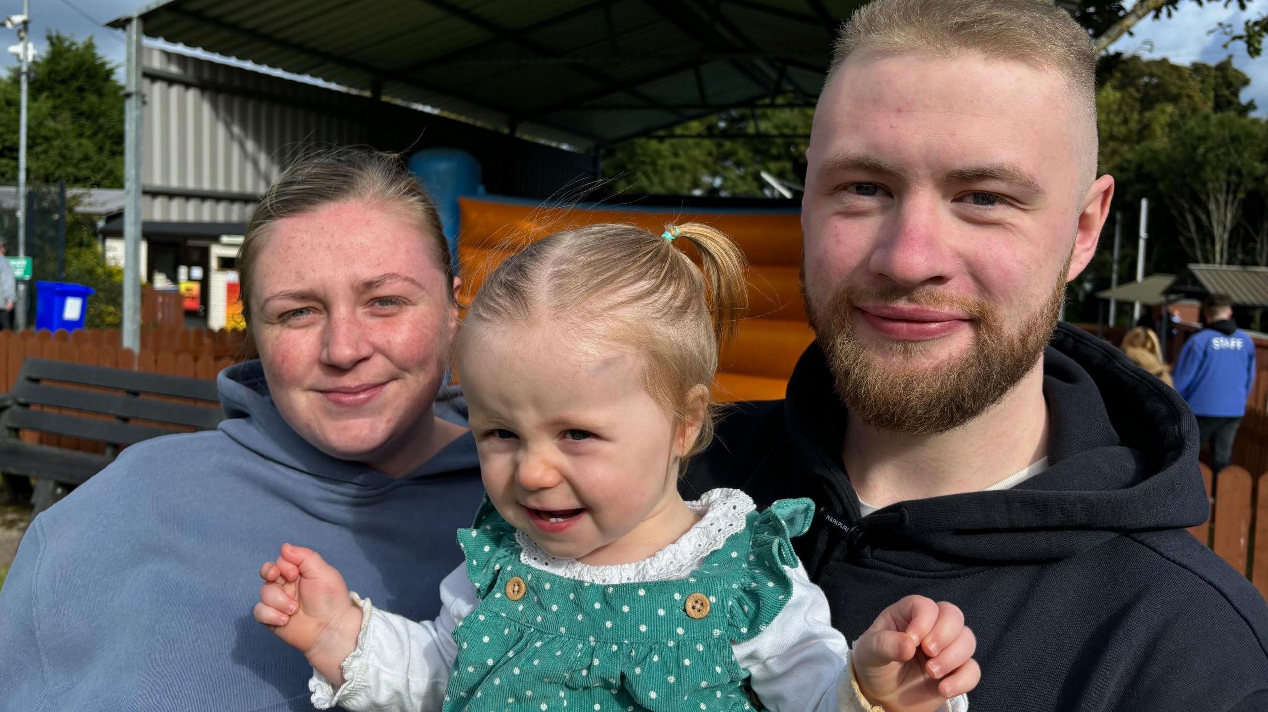 A mother, father and their daughter smile at the camera while attending the fun park at Carnfunnock Country Park. Jennifer is wearing a blue hooded top, their young daughter is earing a white top and green dress and her hair is in pigtails. Her dad has blond hair and a beard and is wearing a black hooded top