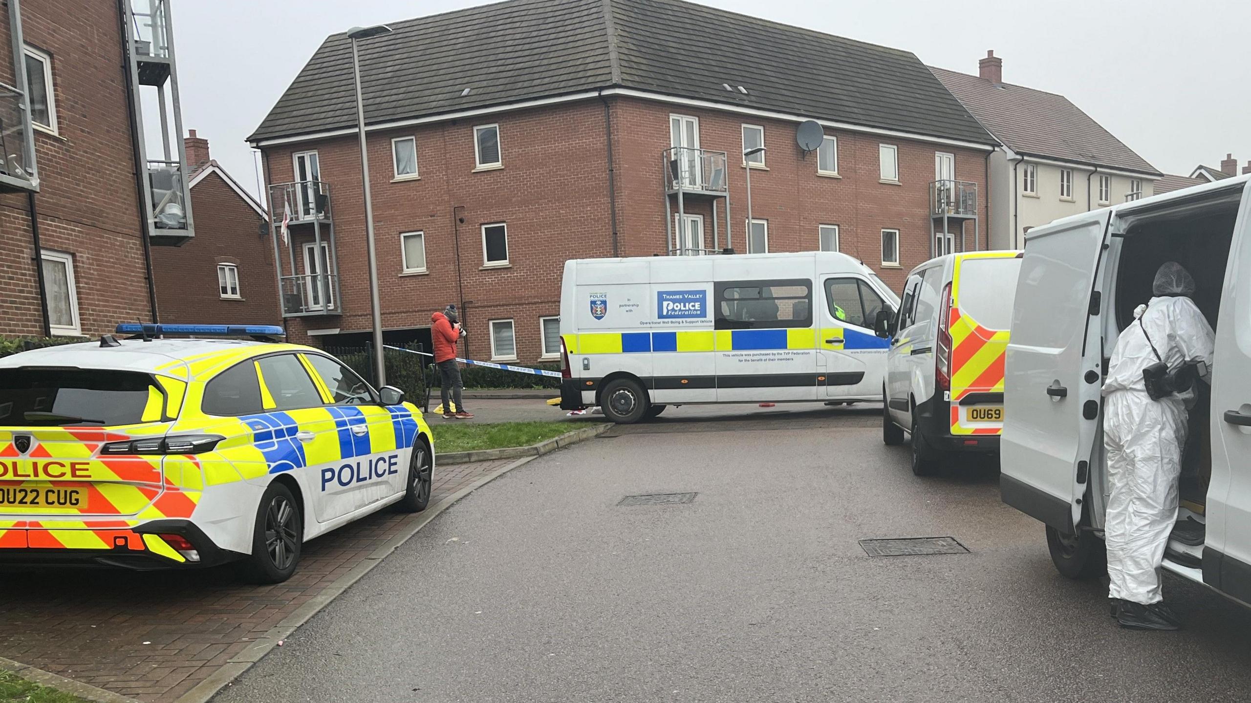 A police car and two police vans, as well as a white forensic van, are parked on the side of a residential road. A forensic officer in a white suit looks into the side of the white van. A police cordon has been placed behind the vehicles. Behind this are several blocks of flats.