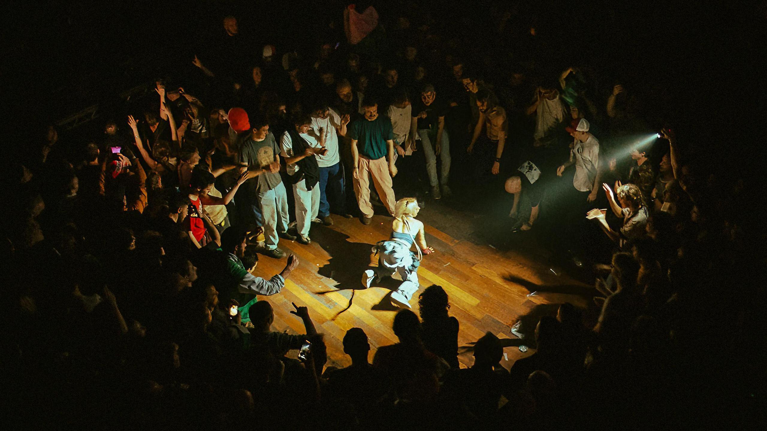 The photo shows a performance from above. A crowd is gathered around a woman who is dancing on the floor. Someone is shining a phone torch on her.