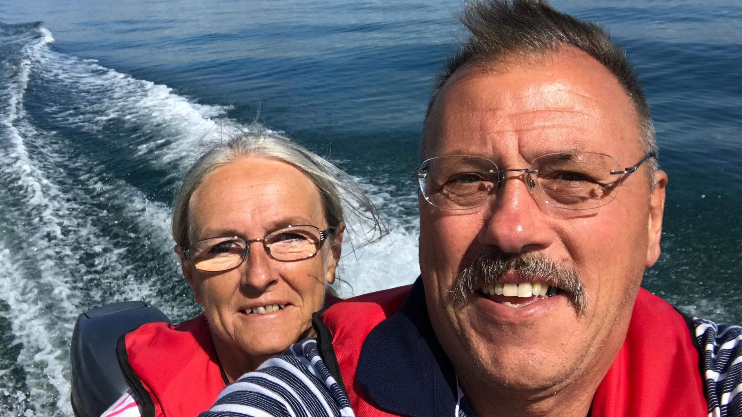 Janet and Paul Ainsworth on a boat, with water in the background. They are both wearing glasses and red life jackets. 