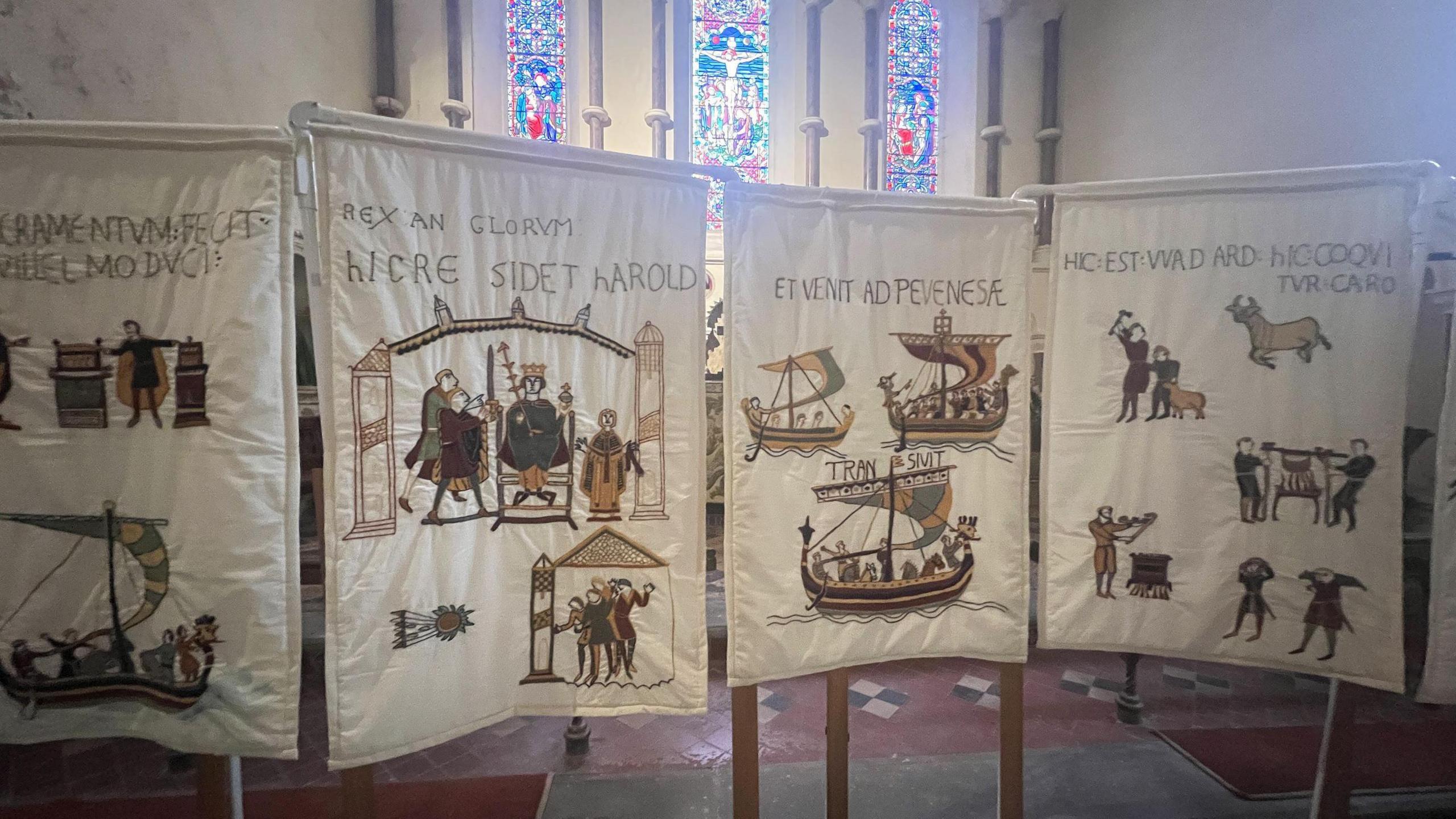 Several quilts displayed like banners are lined up next to each other inside the parish church of St Andrew's, Stogursey, made to look like the Bayeux Tapestry. Sunlight streams through a stain glass window in the background.