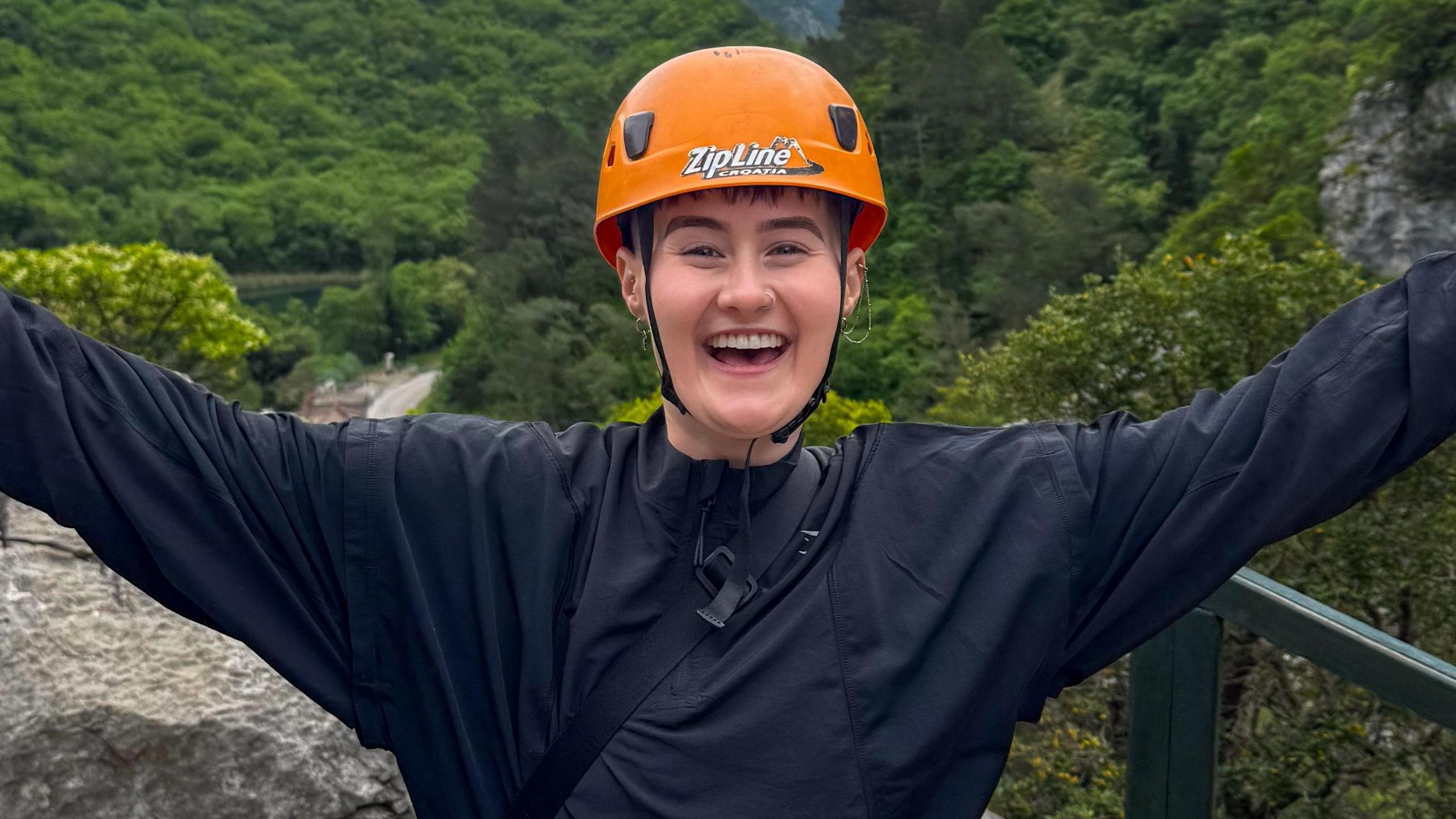 Shell Rowe standing outside. She is wearing an orange helmet which says ZipLine on it in white writing and is wearing a long sleeved black jacket which is zipped up all the way. She is looking at the camera and smiling and has her arms stretched outwards. 