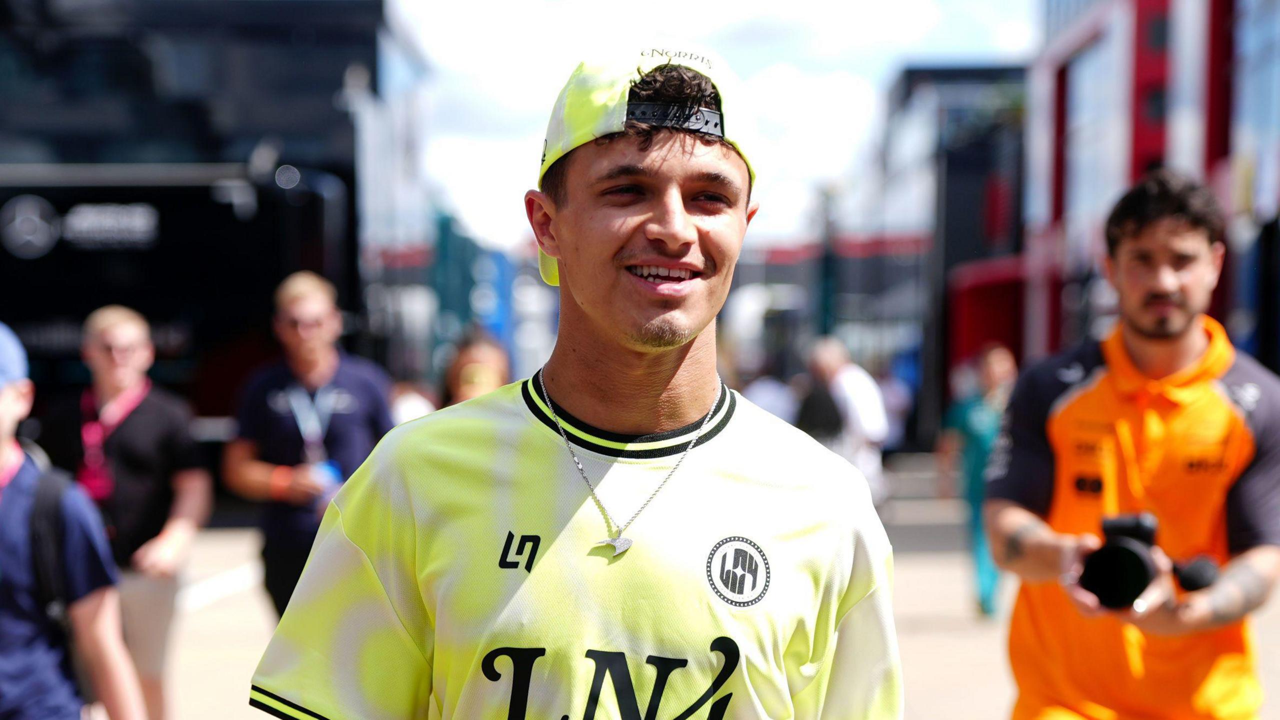 Lando Norris is in the middle looking just off centre. He has a yellow baseball cap on backwards and is wearing a matching yellow and black T-shirt. People are walking behind him in the background.