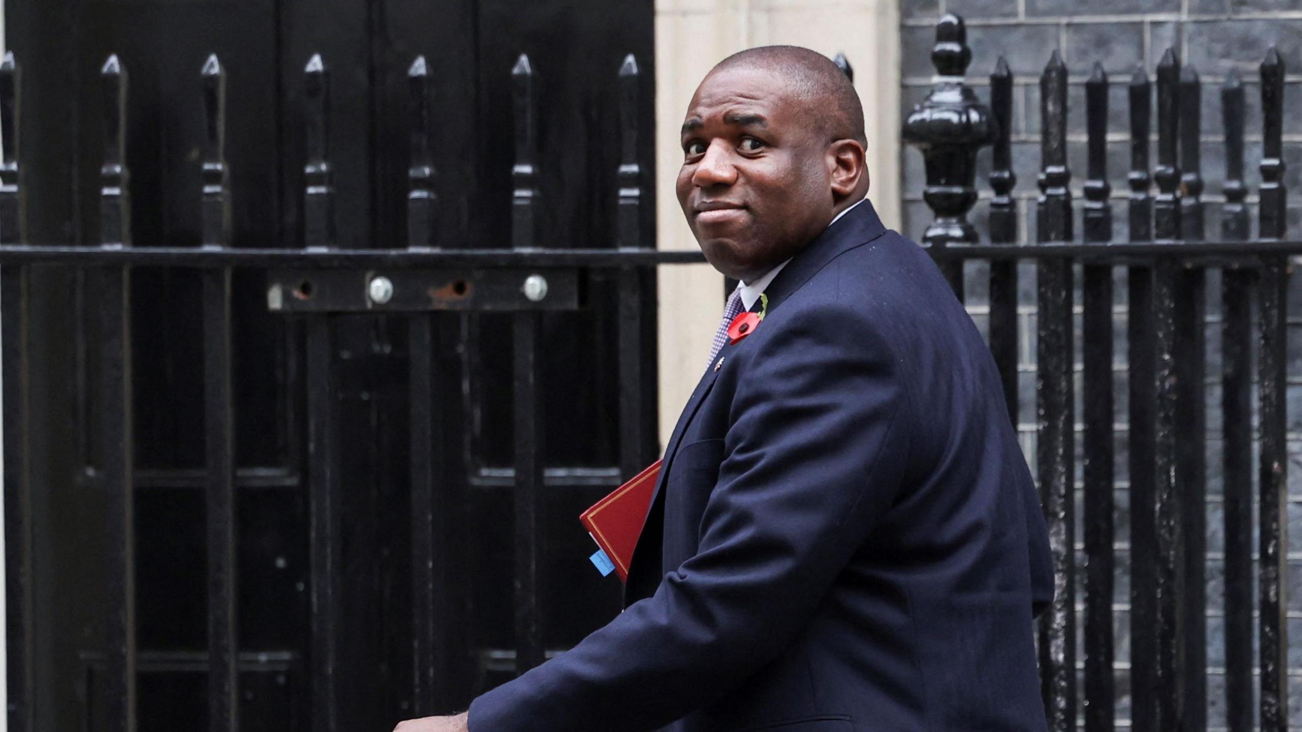 David Lammy entering Downing Street. He is looking back over his left shoulder and is smiling. He is wearing a dark blue suit and is carrying a red ministerial folder.