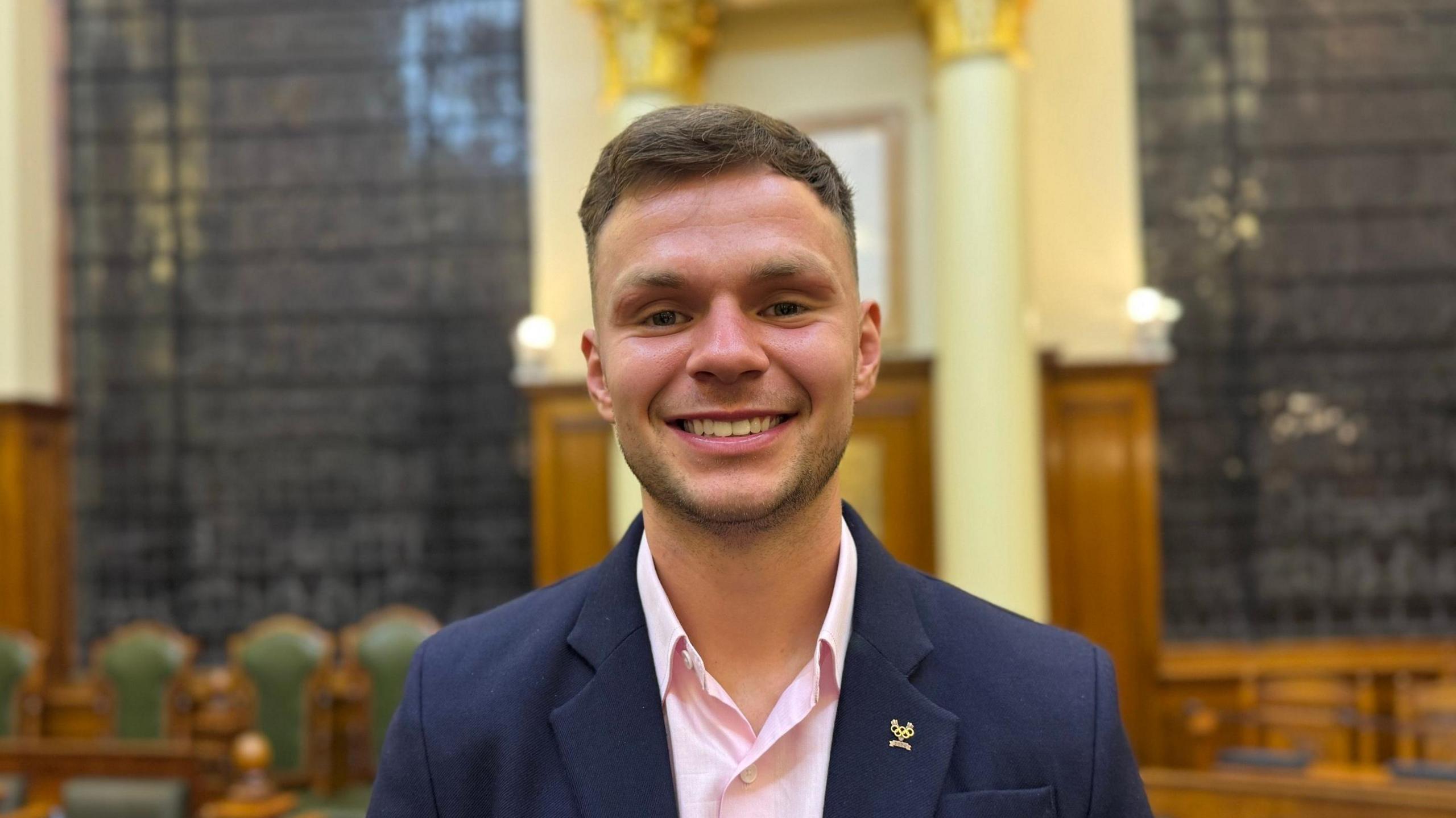 A man smiling straight at the camera. He is wearing a navy suit jacket with a pink shirt and a gold badge featuring the Olympic rings on his lapel.