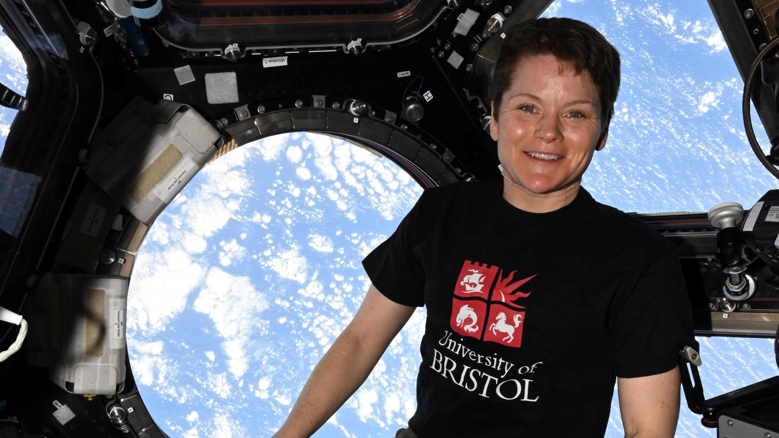 A woman hovers in part of the International Space Station, with part of the earth visible behind her. She is wearing a black T-shirt with "University of Bristol" in white letters on it, below a red and white version of the university's crest