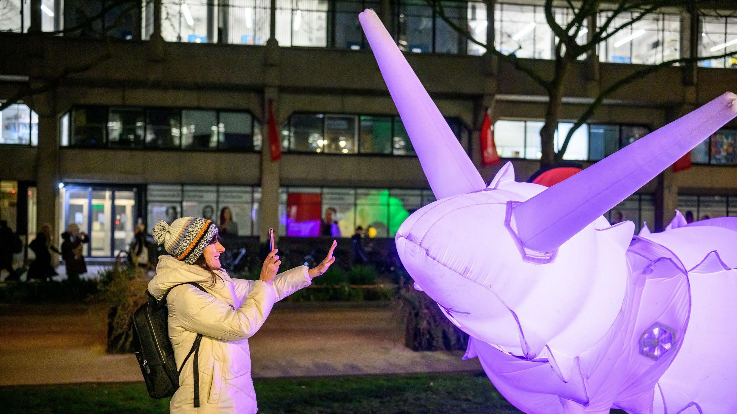 A woman in a bobble hat and white coat smiles as she takes a picture in front of the illuminated snail at night. They are in a public area with buildings behind.