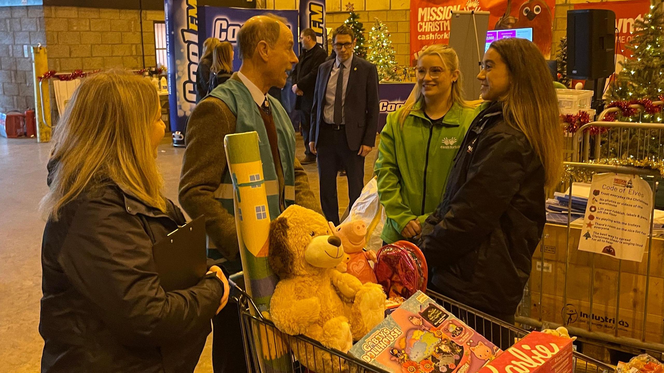 Prince Edward wearing a blue hi-vis as he wheels a trolley of children's toys. He is surround by three females, the first lady has long blonde hair and wears a black coat, the second lady with long blonde hair wears a green Cash for Kids coat and the third has brown hair and wears a black coat.