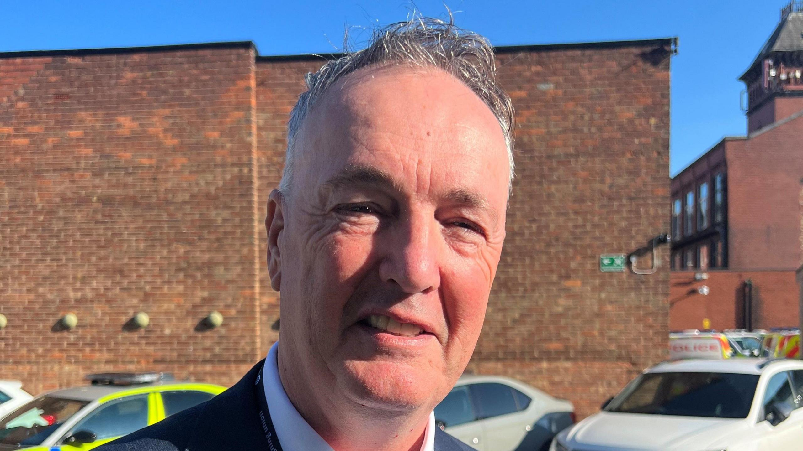 Clive Grunshaw, who has grey hair and is wearing smart clothes, is smiling at the camera. There are police vehicles in the background