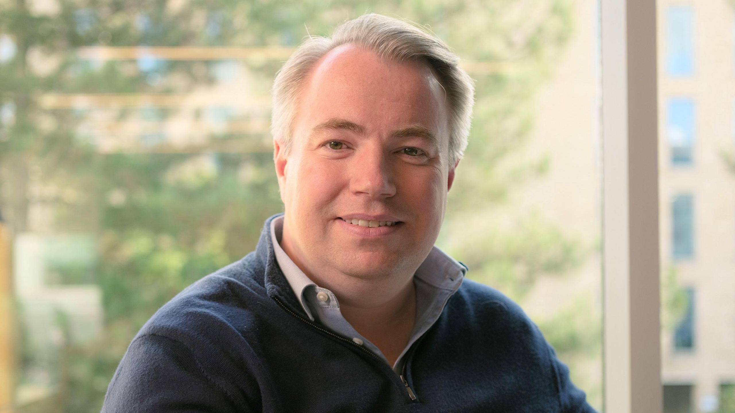 Will Gallagher, sitting down, looking at the camera. He is wearing a light blue shirt, a black blue jumper, has great short hair and is smiling. 