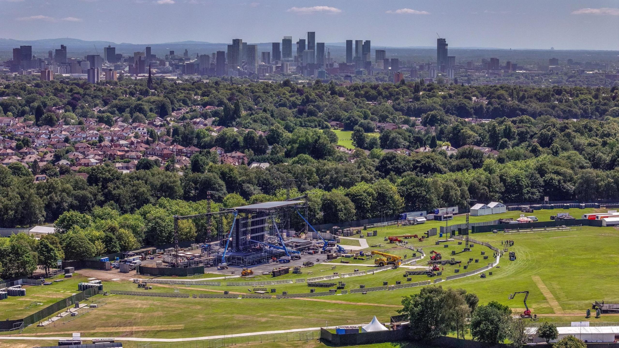 Aerial drone view of stage being built at Heaton Park surrounded by construction vehicles in front of rows of trees. In the backdrop are residential houses plus high-rise buildings of Manchester city centre. Pennine hills can be seen in the background.