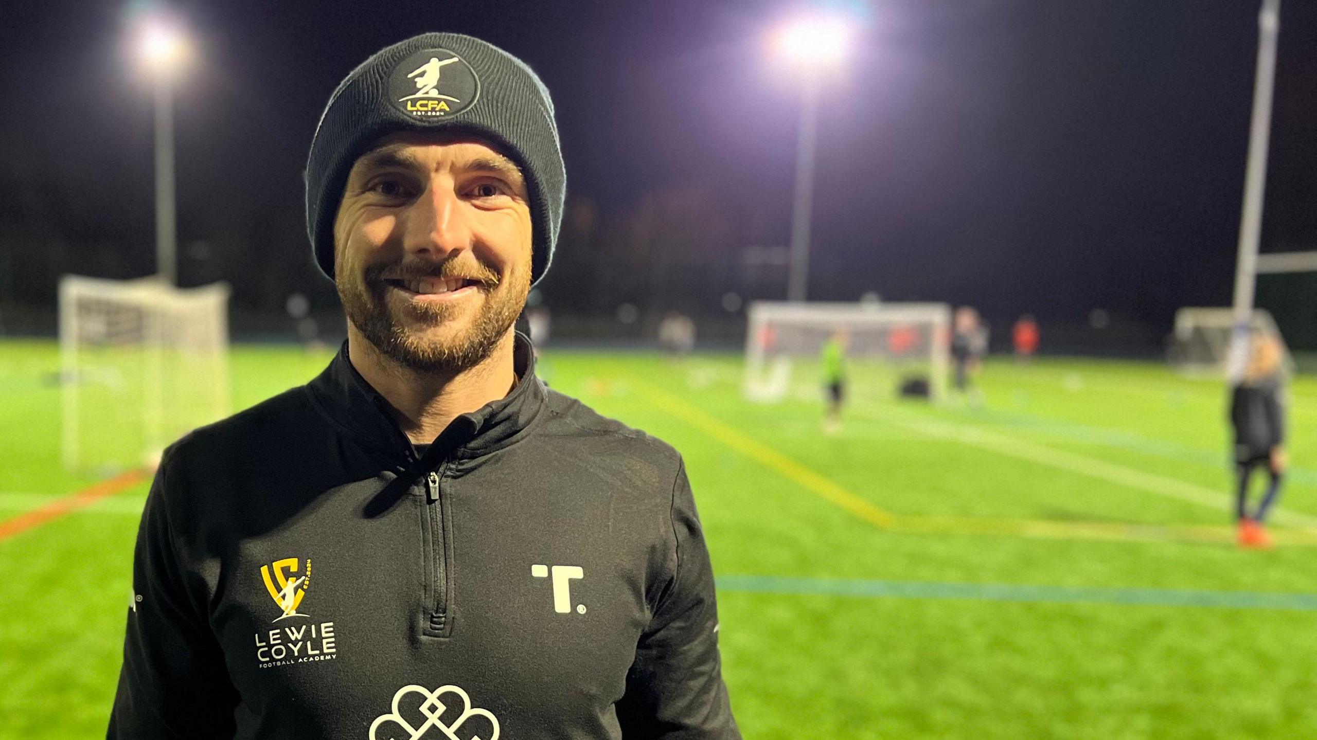 Lewie Coyle, a professional footballer, stands on a floodlit football pitch as children play in the background. He is wearing a black beanie hat and a black sports top displaying the logo of his football academy.