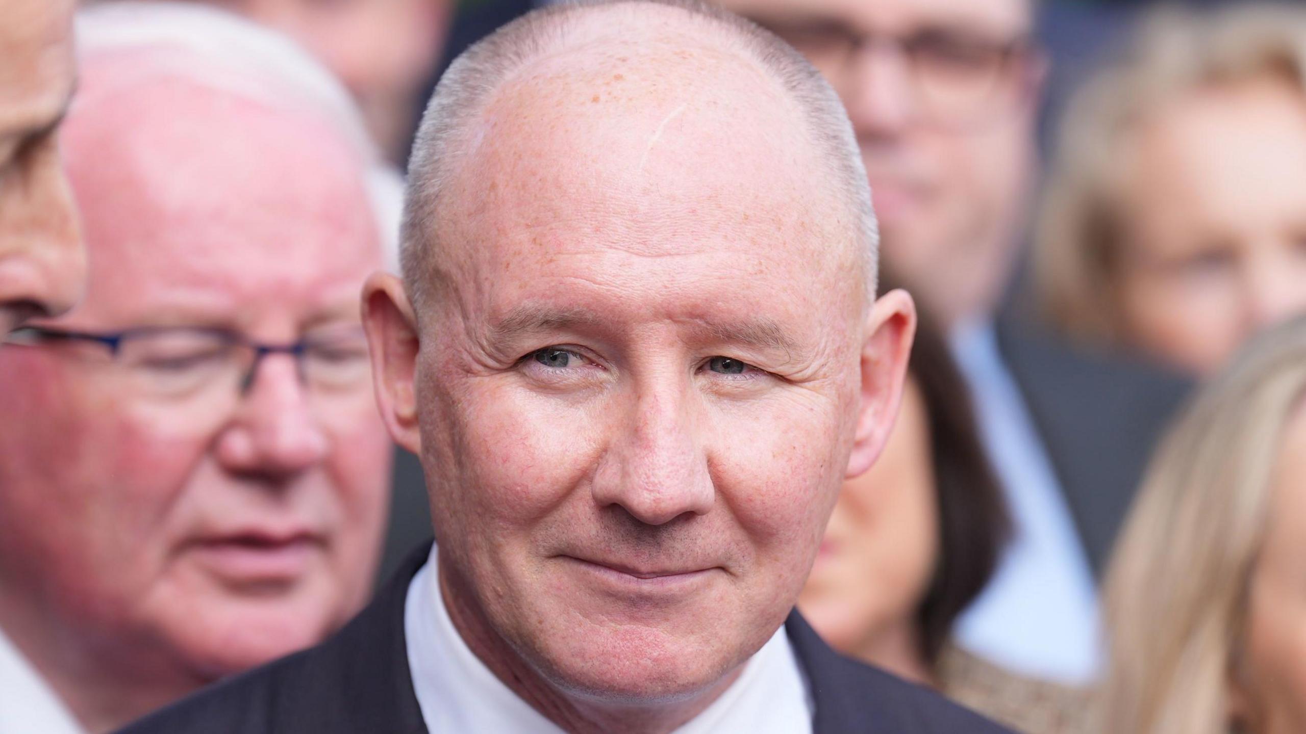 Jim Gavin wears a black suit, white shirt and dark blue tie. He is in a crowd of people.