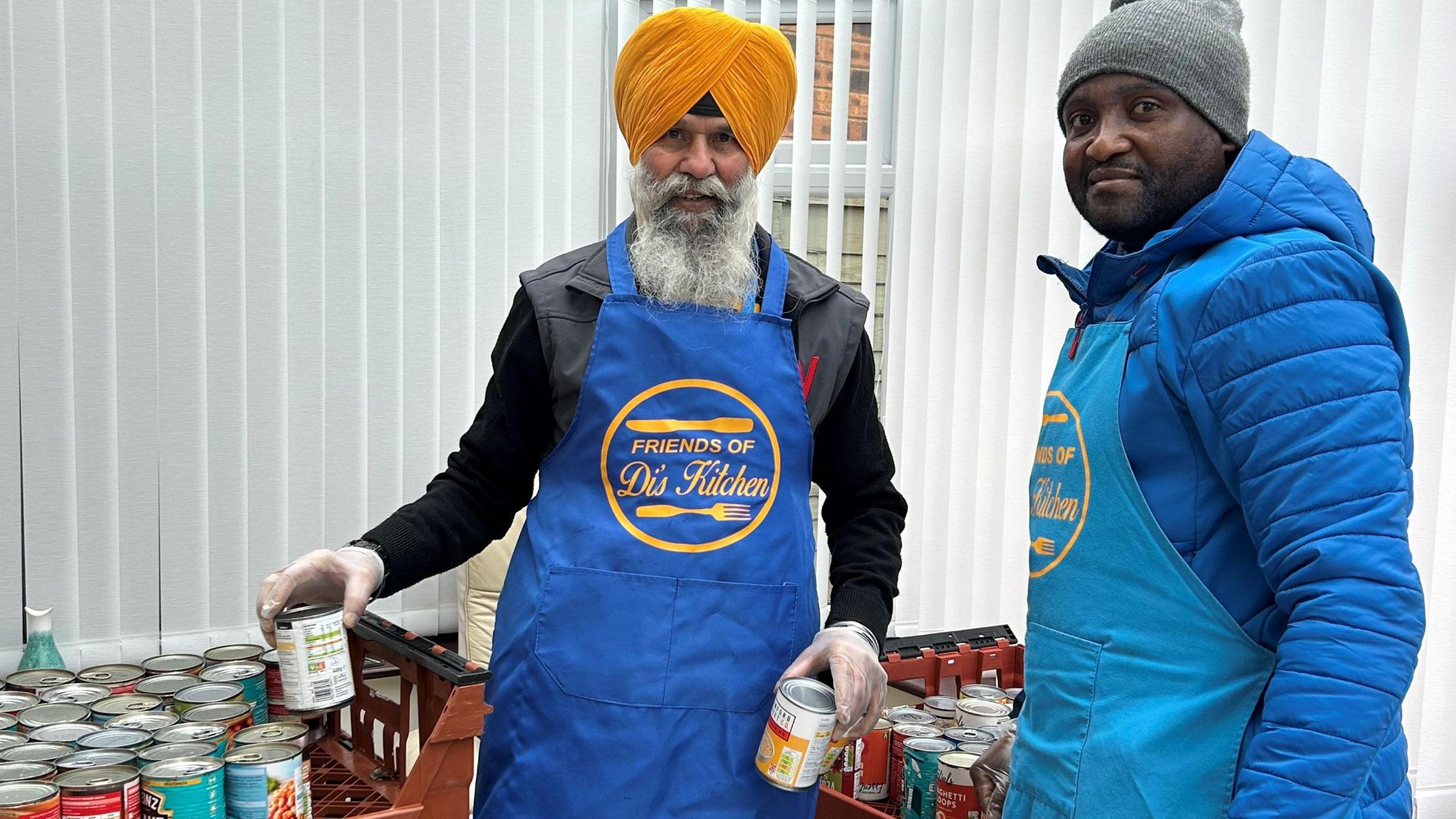 A man wearing a turban with a grey beard holds tins of food in his hands. Another man wearing a hat stands next to him. Both are wearing blue "Friends of Di's Kitchen" aprons. 