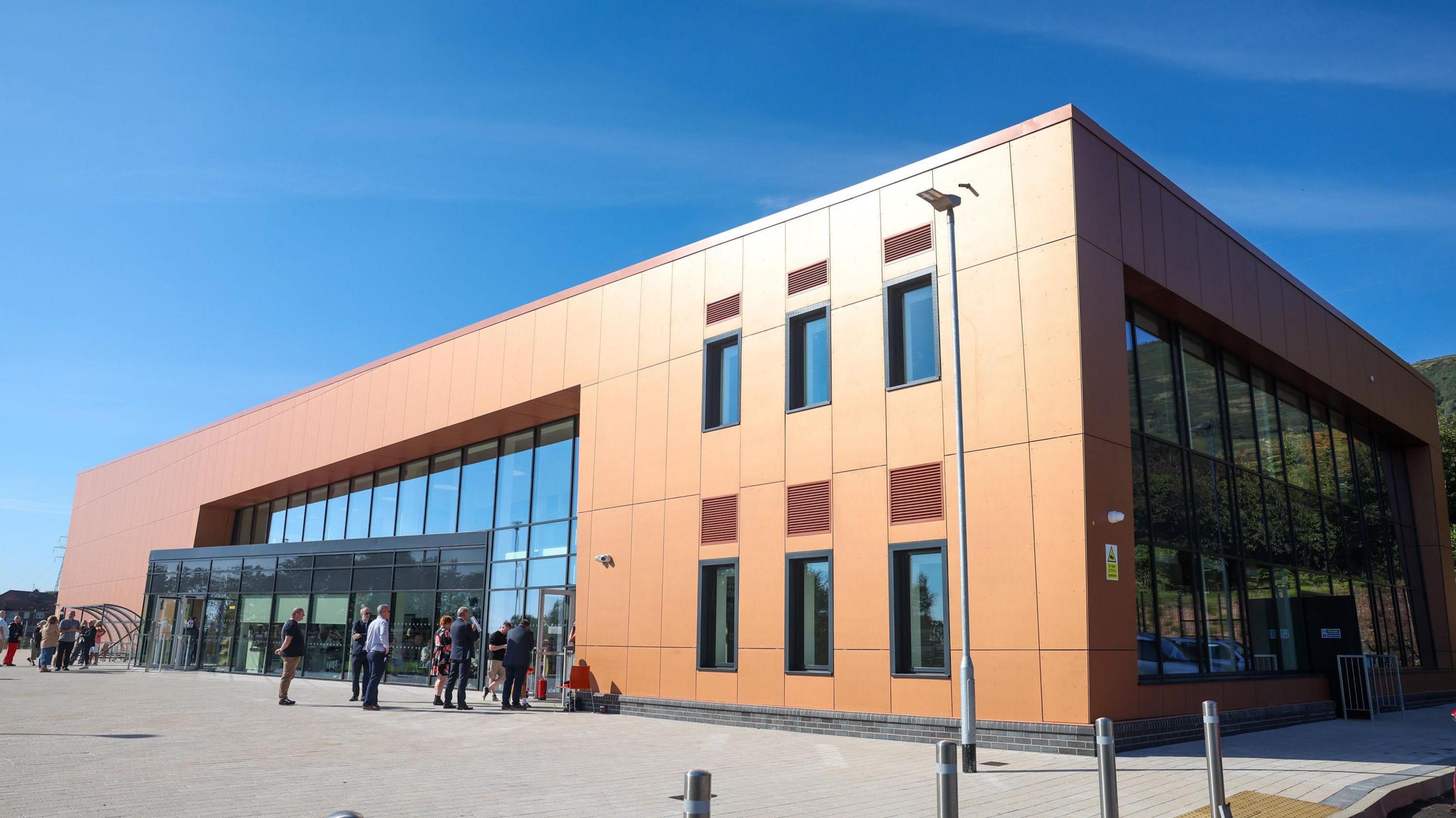 The outside of the Black Mountain Shared Space building. It is a large boxy building with copper cladding and large windows. There is a blue sky with no clouds in the background. 