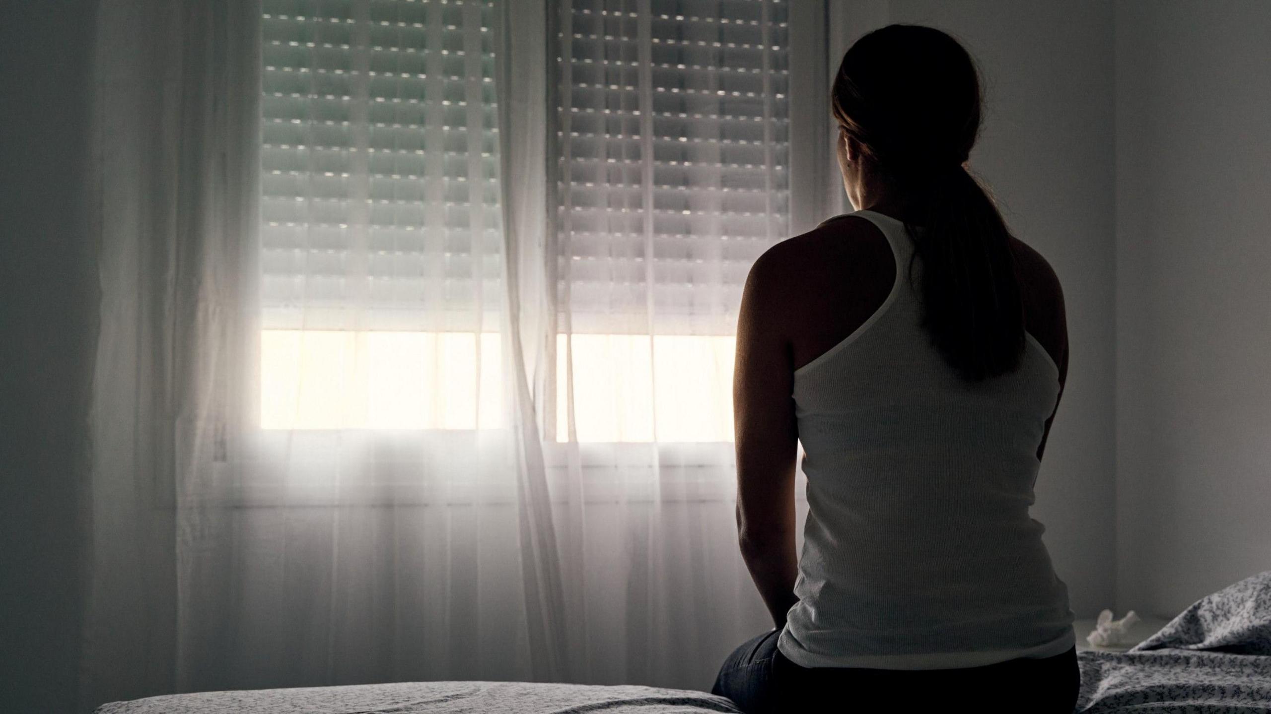 A woman with long brown hair in a ponytail is sat on the edge of the bed facing a white wall and window.