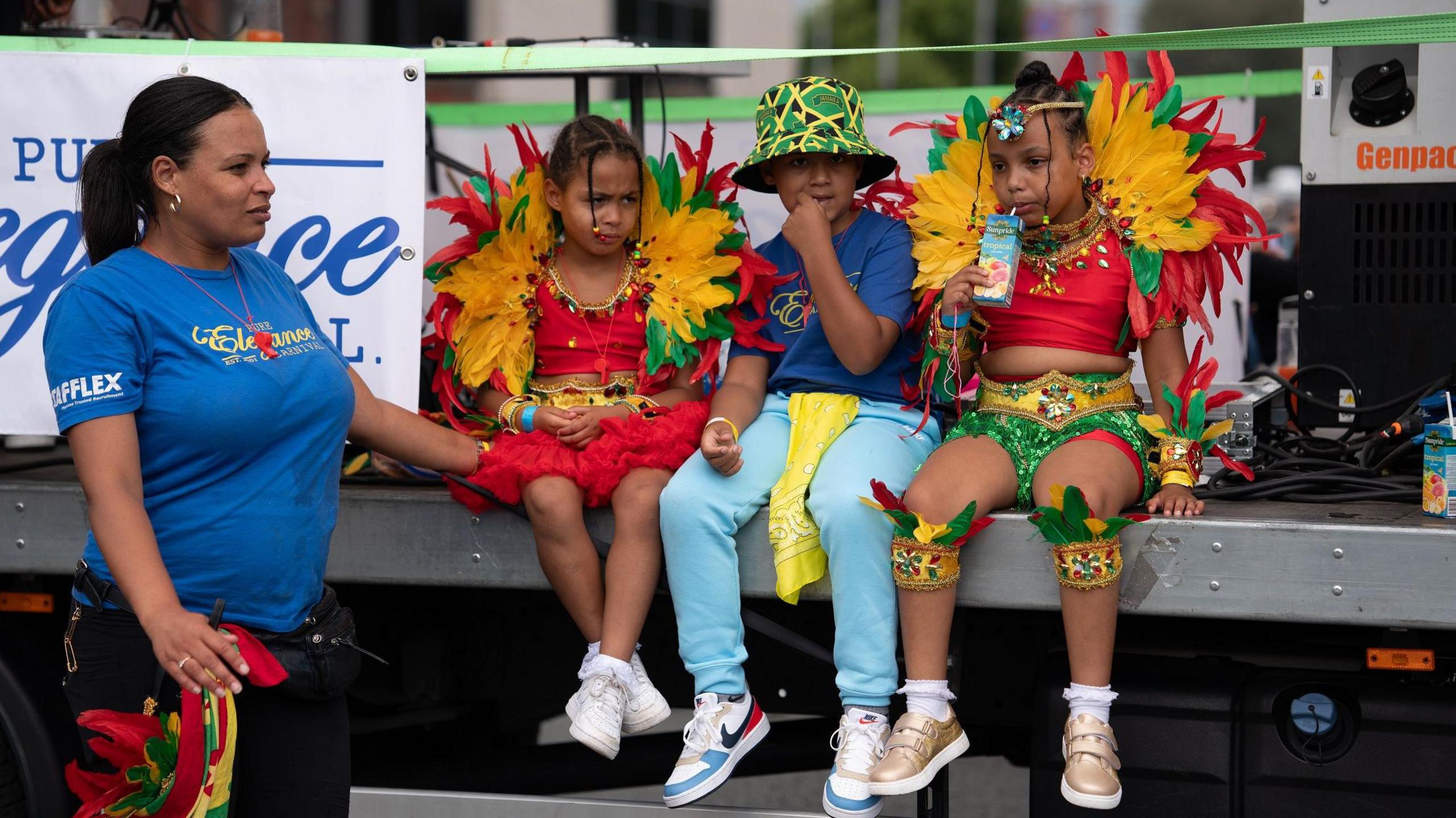 Three children - a boy flanked by two girls in red dresses - sit on a stage above the ground. The girls are wearing yellow, green and red feathers around their heads and shoulders as one of them sips from a juice carton. The boy is wearing a bucket hat in the yellow, black and green design of the Jamaican flag. A woman who is wearing a blue sleeveless top stands to the side, keeping an eye over them.