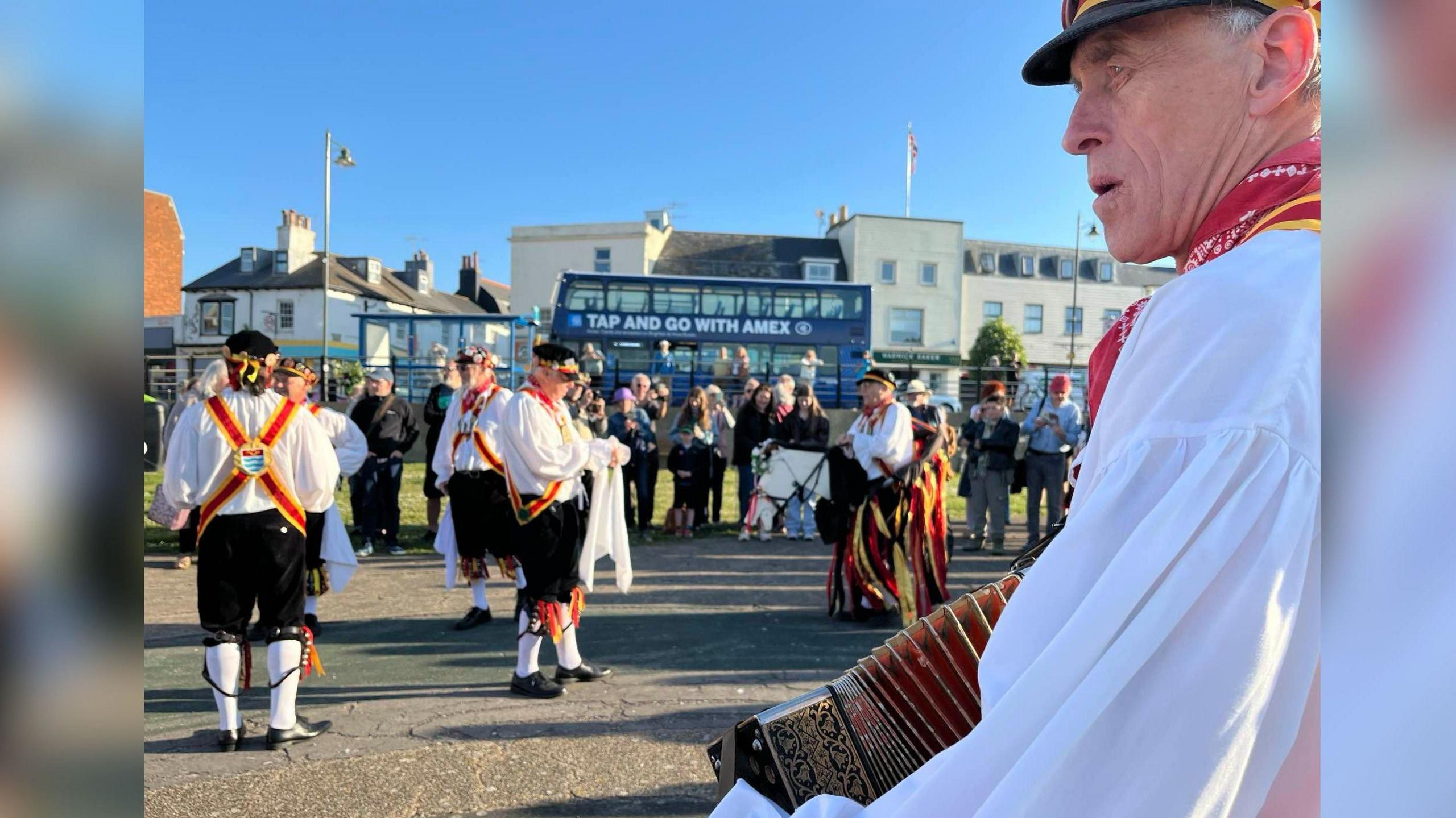 A group of Morris dancers seen in their outfits of white shirts and black trousers with yellow and red ribbons and bells on their legs. In the foreground is a man seen from the side holding an accordion.