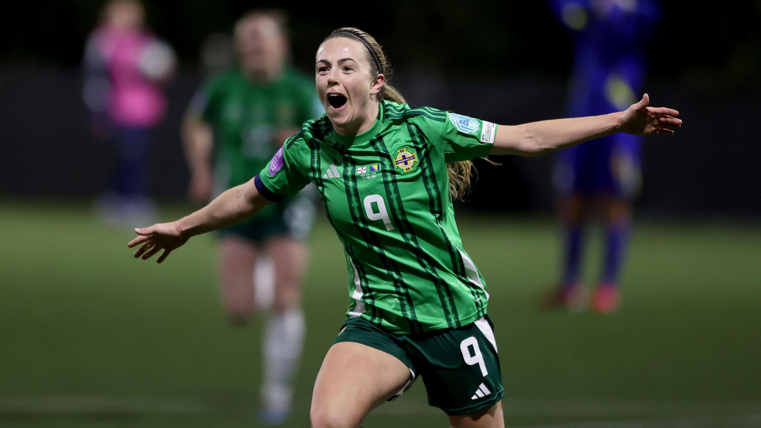 Northern Ireland's Simone Magill holds her arms out in celebration after scoring a goal. She is wearing dark green shorts with the number 9 on them. Her top is a lighter green with some darker green stripes. It also adorns the number 9 on the chest