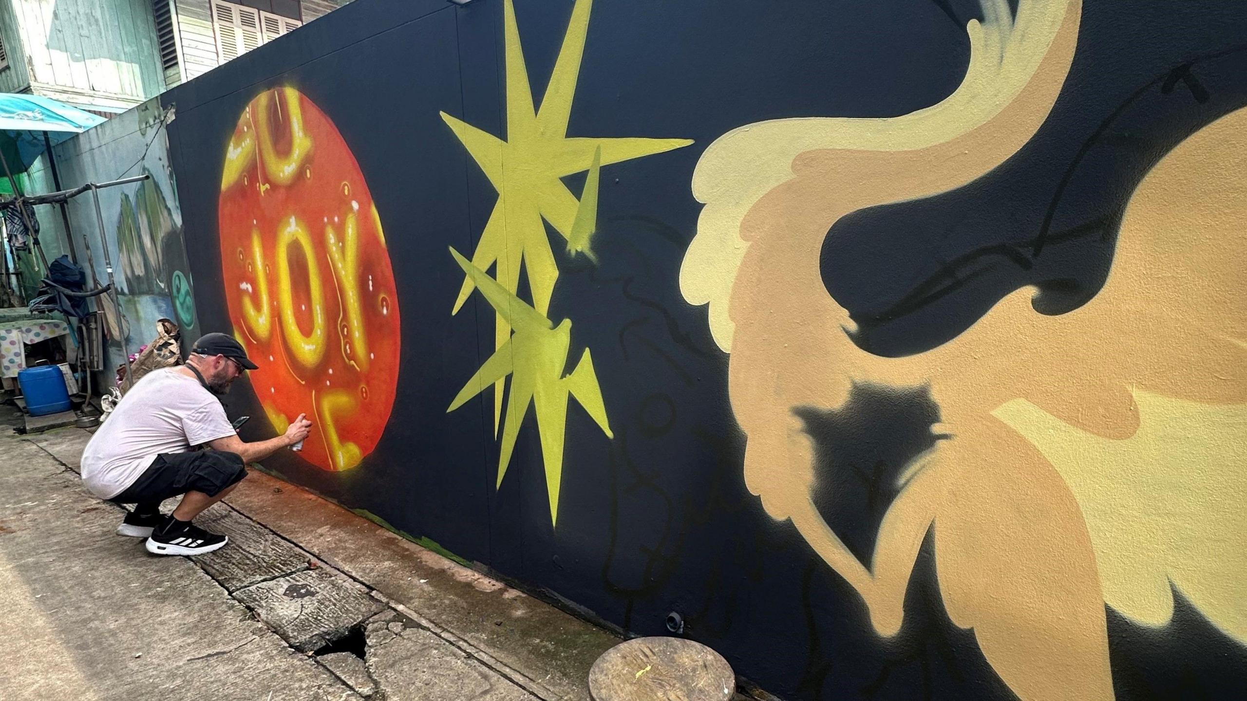 A man in a white top, black shorts, black runners and black hat - is crouching as he holds a spray can which he is using to paint a mural on a dark wall. His mural is of alphabet spaghetti which spells joy. A mural of stars and a swan can be seen beside it.