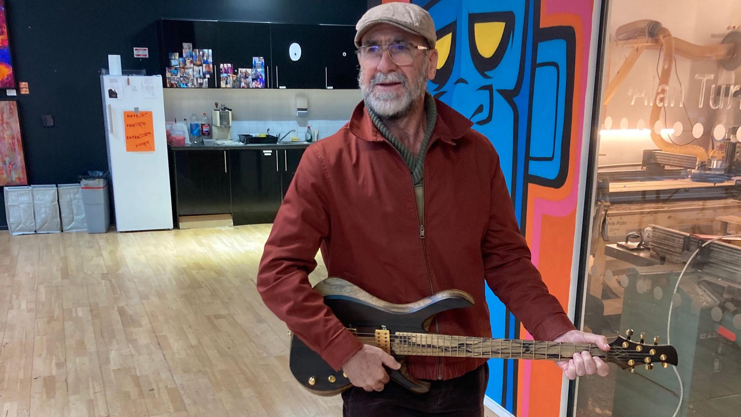 Eric Cantona is playing a wooden guitar. He is wearing a red jacket and a flat cap. 