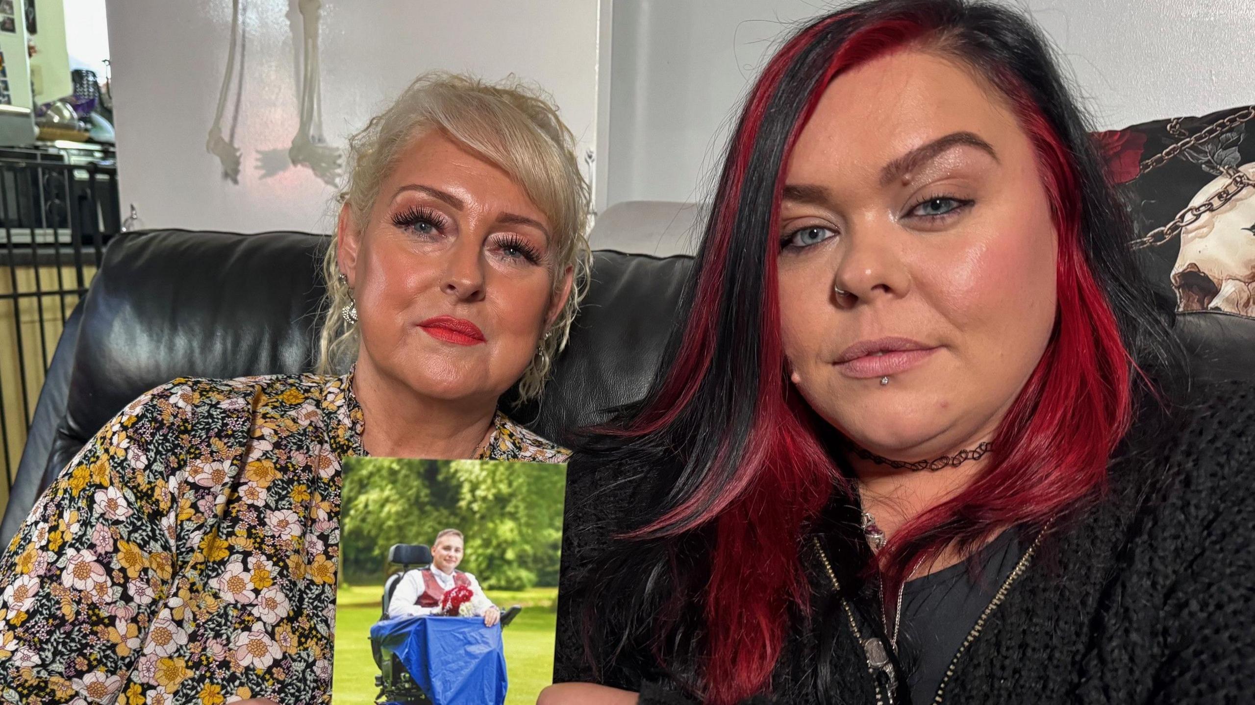 Tracy Lee, a woman in her fifties wearing heavy make-up and a floral top sitting next to her daughter-in-law Sammie Lee who has black hair with red streaks. They are carrying a photograph of Karl Lee, who is sitting in a wheelchair in the photograph.