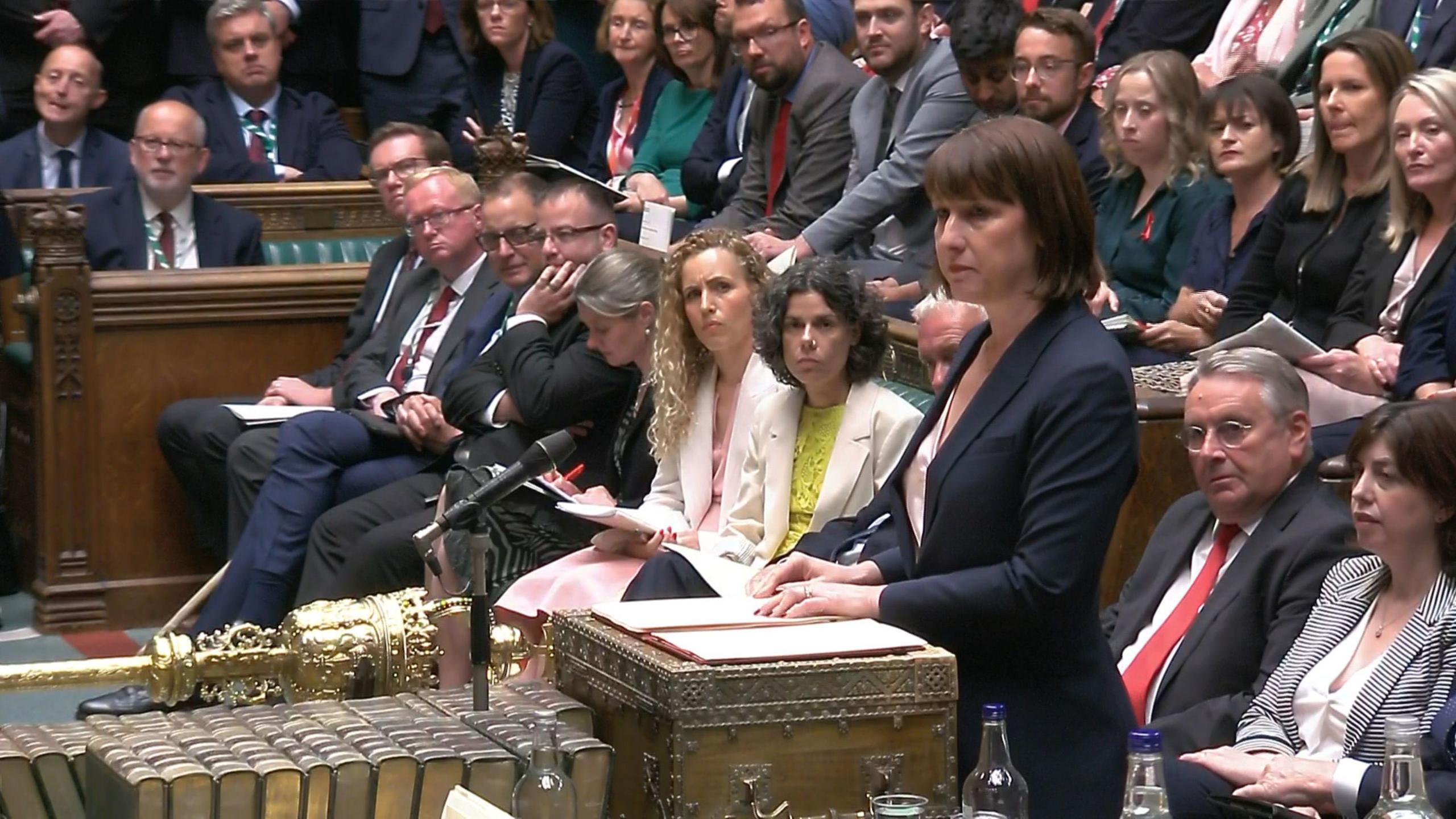 Rachel Reeves standing and looking solemn as she addresses the House of Commons, with the Labour frontbench and MPs seens behind her