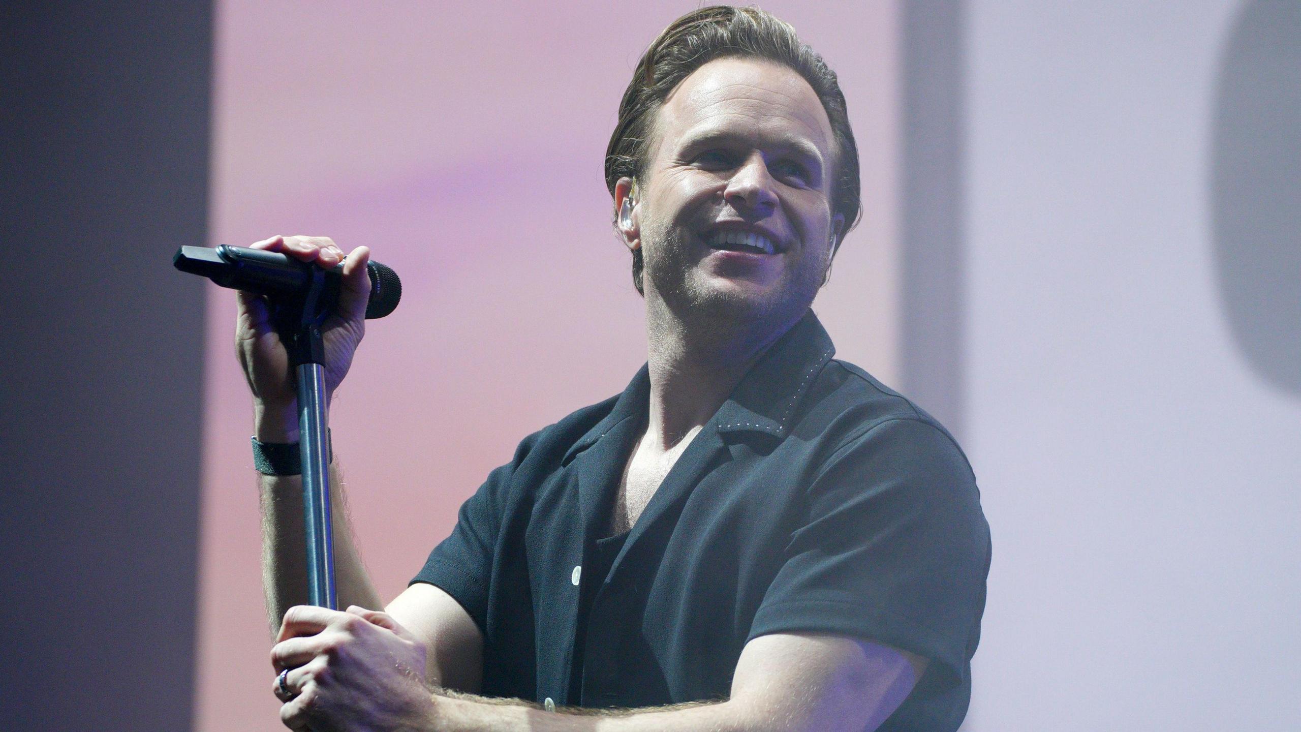 Singer Olly Murs stands at a microphone, smiling over his shoulder.