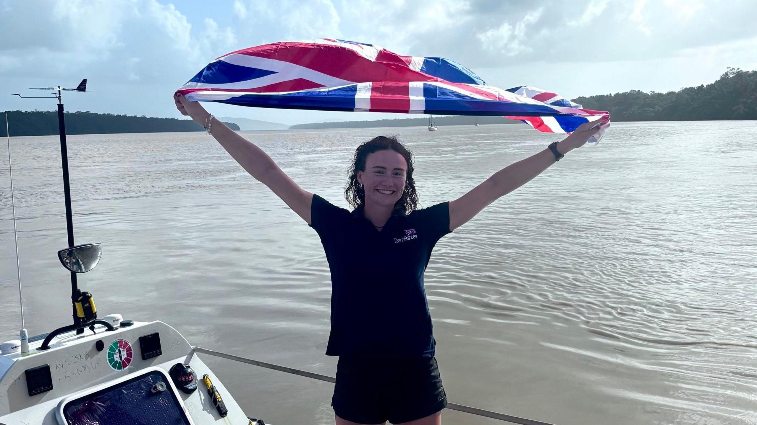 Zara is holding a large Union flag above her head. She is smiling at the camera and wears a blue Team Forces branded polo shirt. Behind her is the sea. Zara has brown curly hair that looks slightly wet at the ends from the rowing.